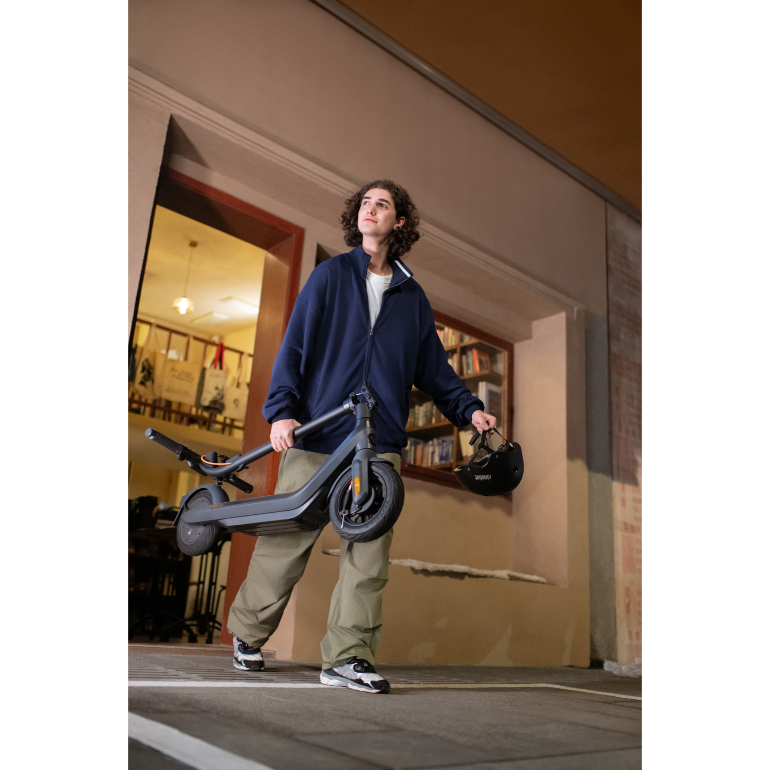 Man carrying a scooter and helmet walking down a street at night.