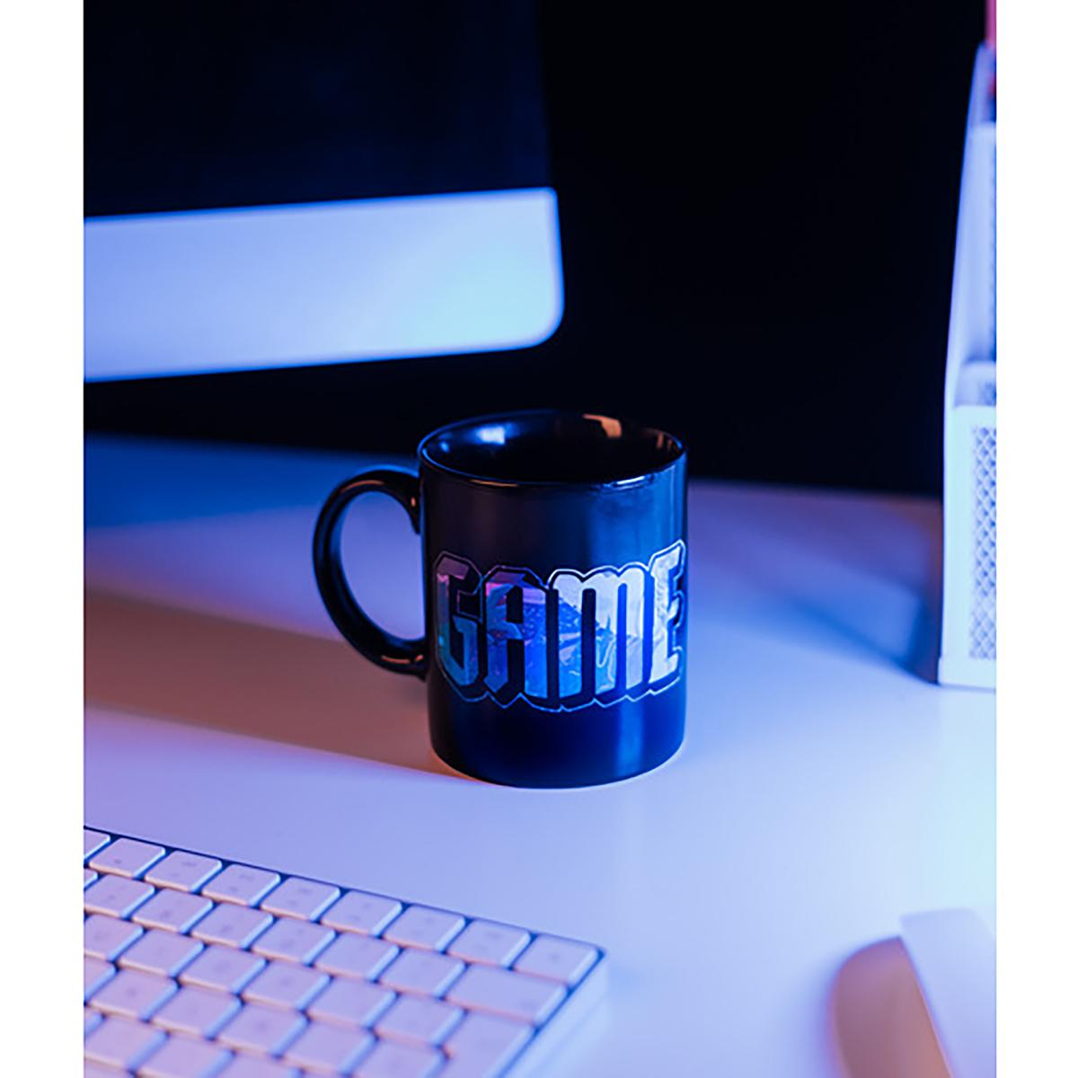 Schwarze Tasse auf einem Schreibtisch mit dem Wort 'GAME' in bunten Buchstaben. Im Hintergrund ein Computerbildschirm.