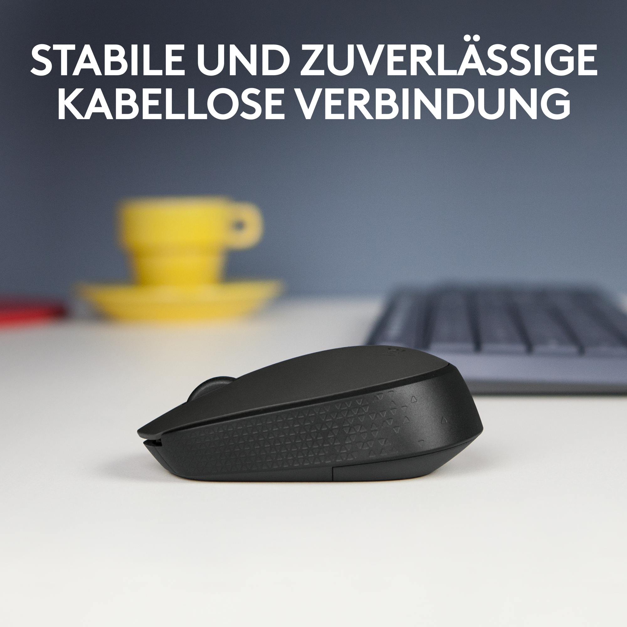 Schwarze kabellose Maus auf weißer Oberfläche. Hintergrund zeigt Tastatur und gelbe Tasse.