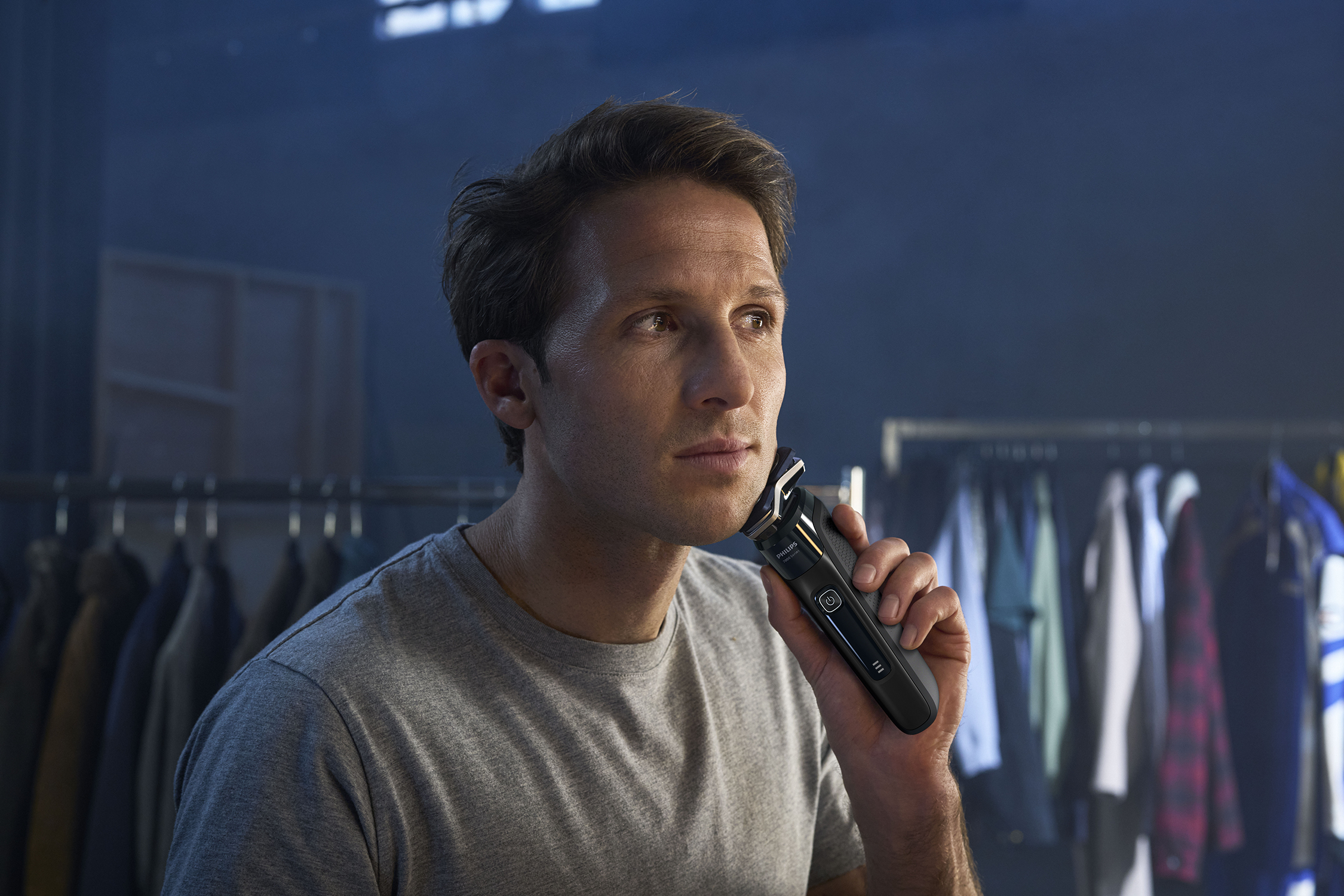 Man shaves with a black electric shaver. Background: clothing rack, mirror, and dark blue wall.