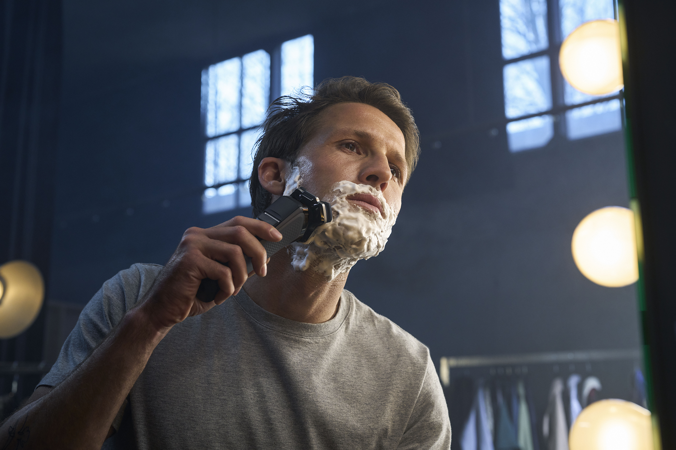A man shaves his face with a black electric shaver, with shaving cream on his face.