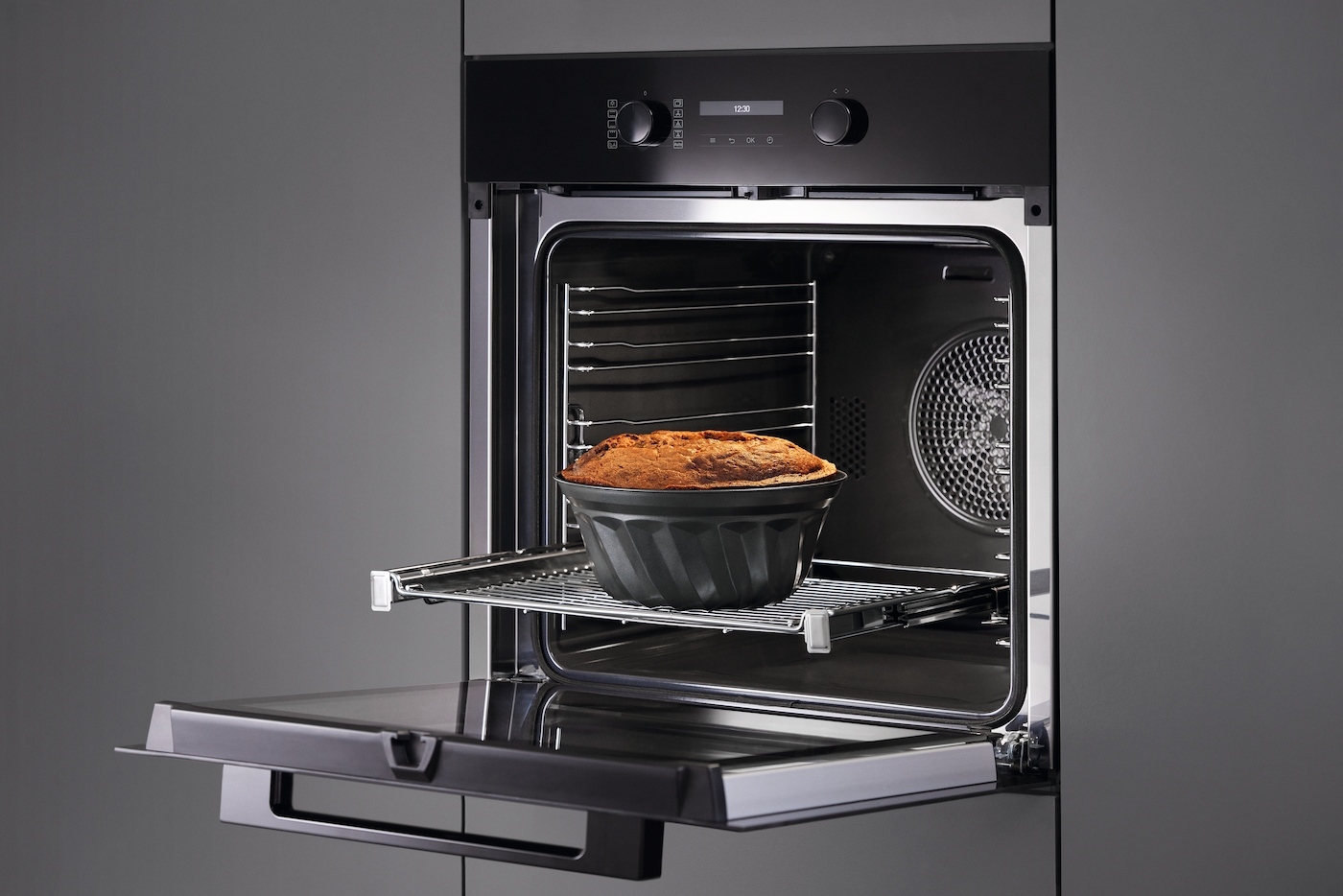 Schwarzer Backofen mit geöffneter Tür, der einen Kuchen in einer dunklen Pfanne auf einem Rost zeigt. Dunkelgrauer Hintergrund.