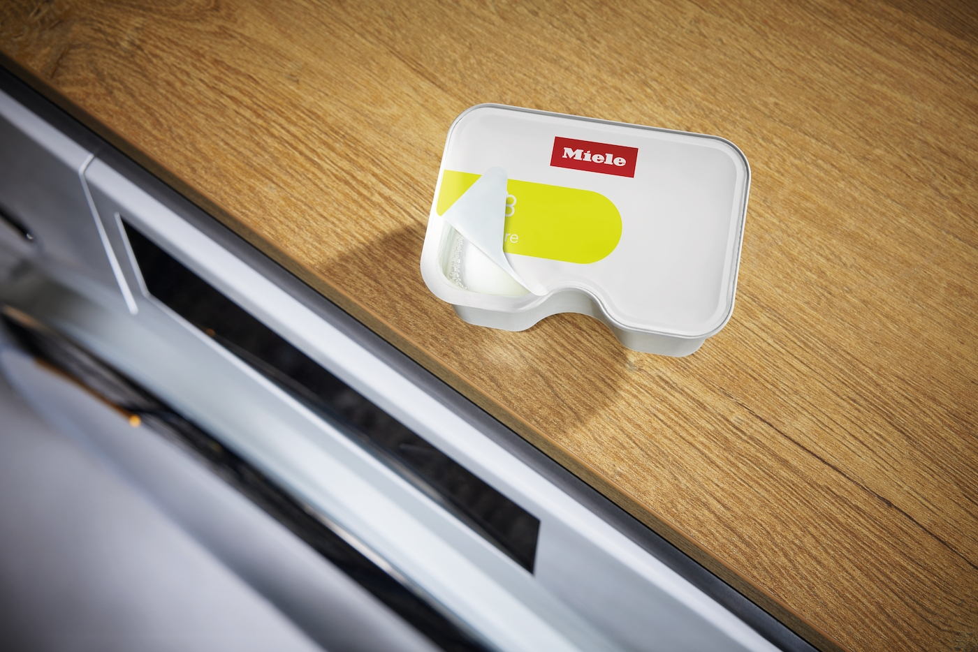 Ein Miele Spülmittelbehälter mit gelbem Etikett auf Holzoberfläche, daneben eine Maschine.