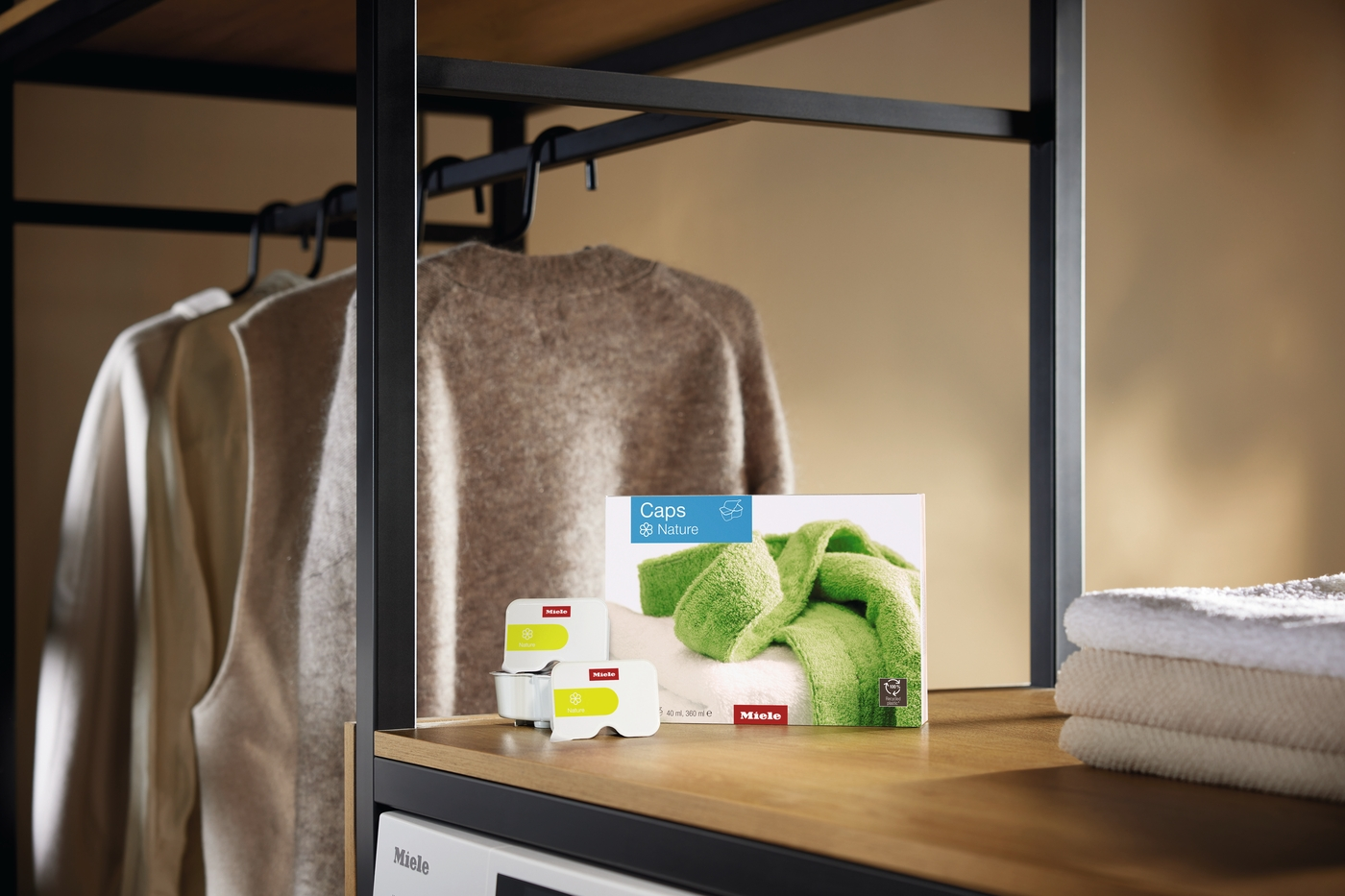Miele Waschmittelkapseln und Kleidung auf einem Regal. Grün und beige.