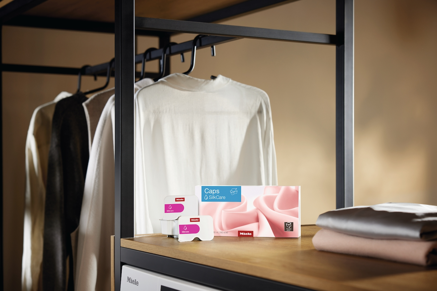 Miele SilkCare Kapseln auf einem Regal neben Kleidung und gefalteten Stoffen.