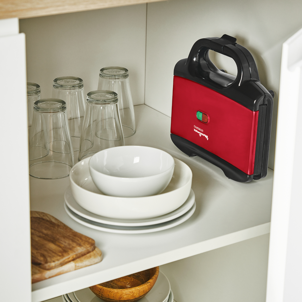 Een rode sandwichmaker met een groen lampje en een handvat, in een witte kast.