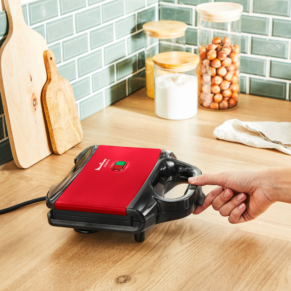 Rode en zwarte Moulinex-sandwichmaker op een houten oppervlak. Een hand reikt naar de handgreep.
