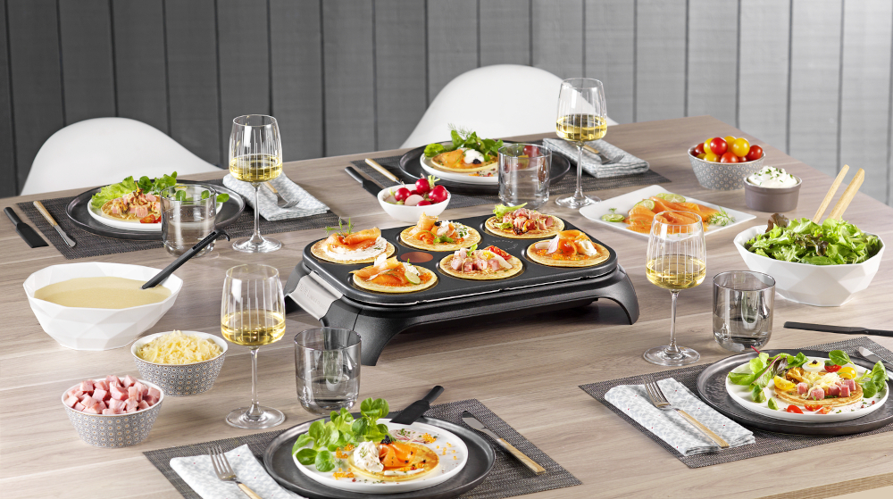 Eettafel gedekt met eten, drinken en een raclette grill in het midden.