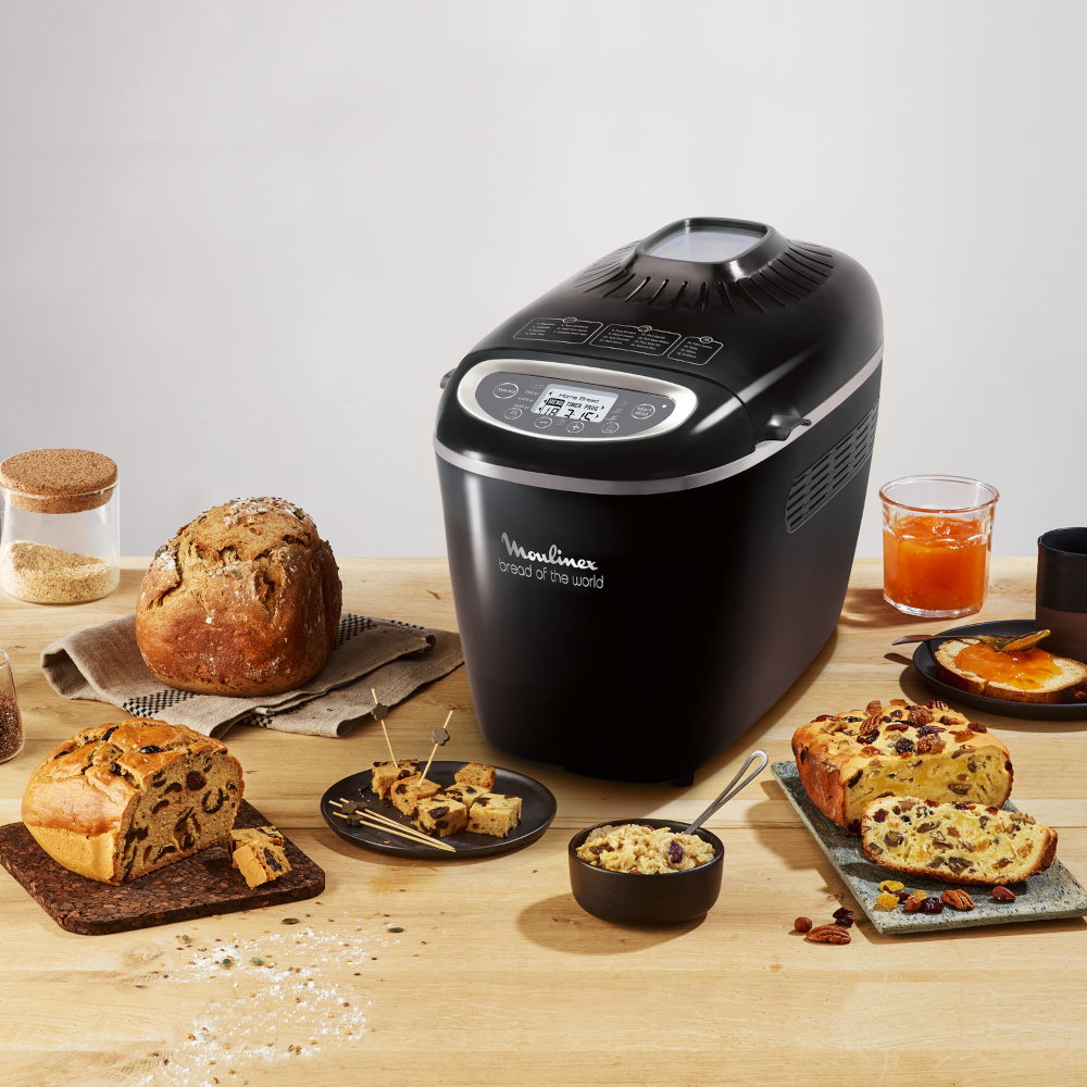A black bread maker with bread and ingredients on a wooden table.