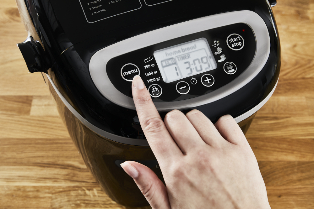 A hand presses the menu button on a black bread maker. The display shows the timer and settings.