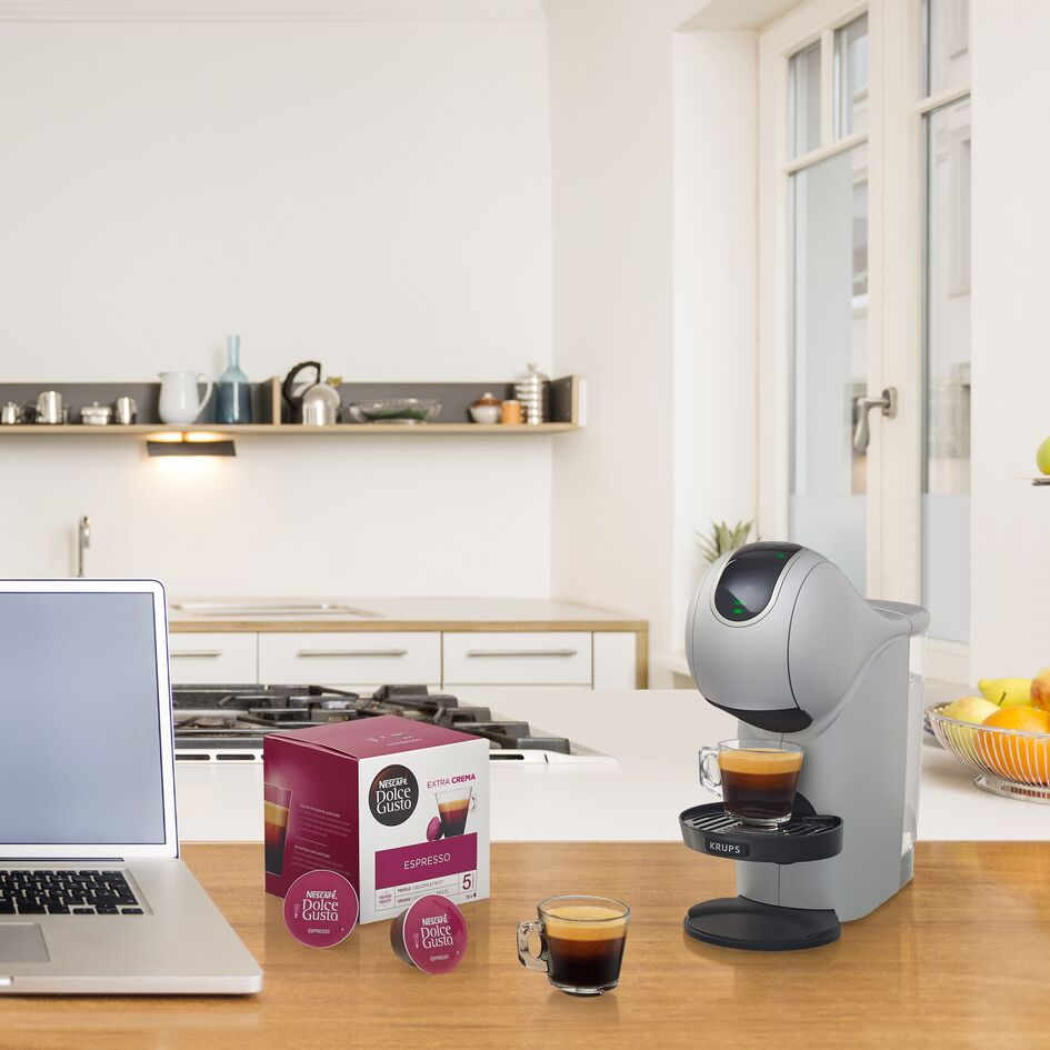 Koffiezetapparaat op een houten tafel, naast koffiepads en een laptop. Keuken op de achtergrond.