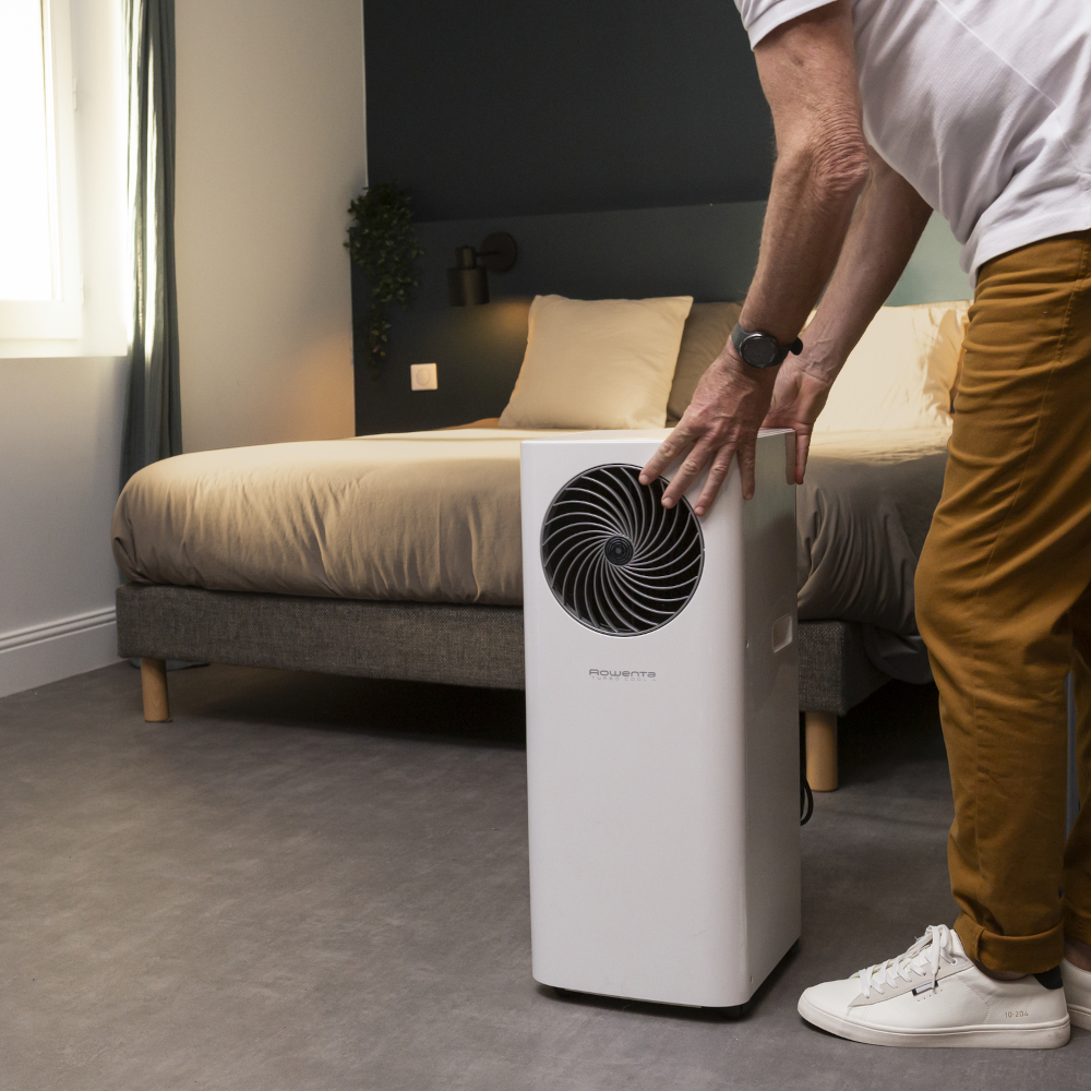 Man naast een witte luchtreiniger in een slaapkamer. Bed en raam op de achtergrond.