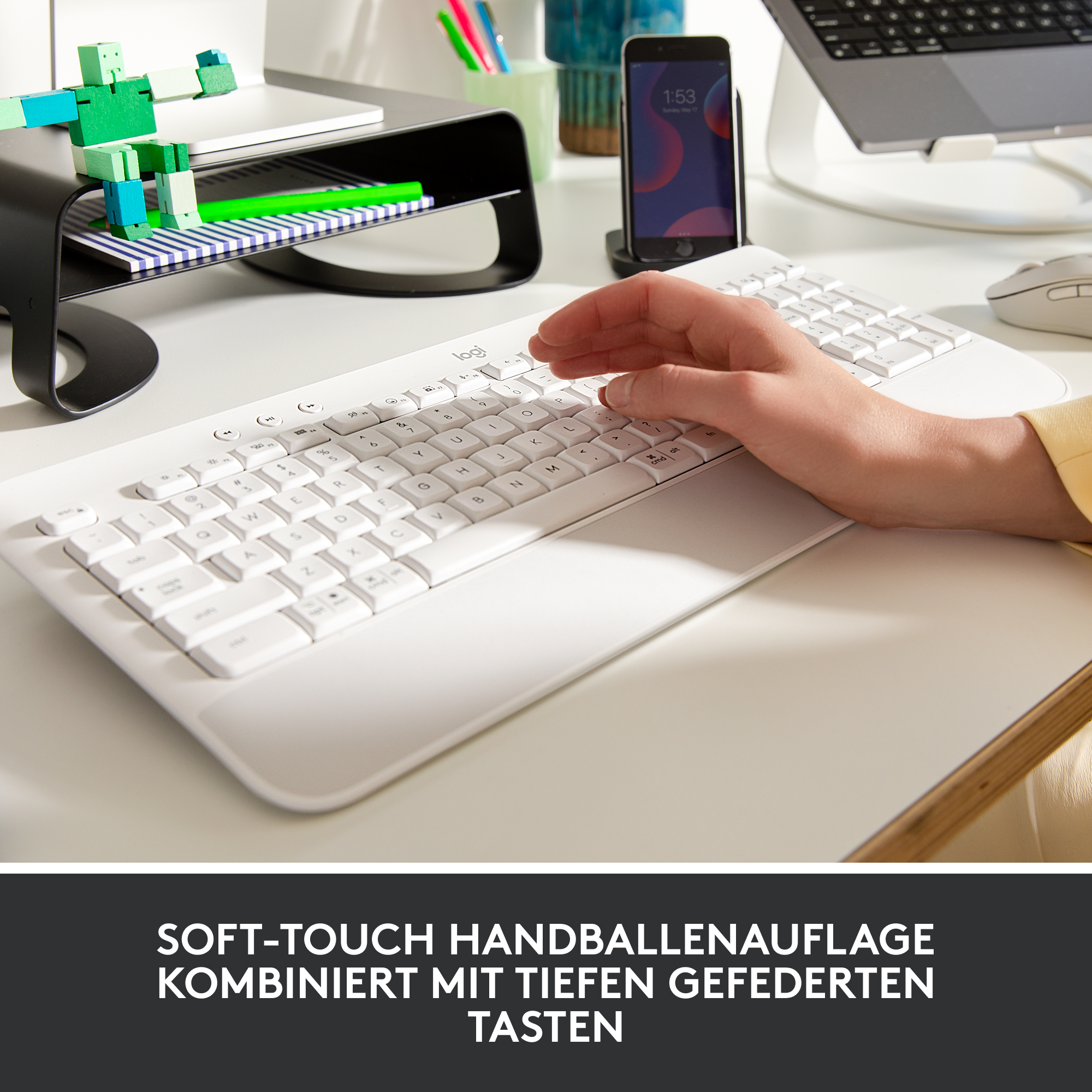 Eine Hand liegt auf einer weißen Tastatur. Weitere Gegenstände liegen im Hintergrund auf dem weißen Schreibtisch.
