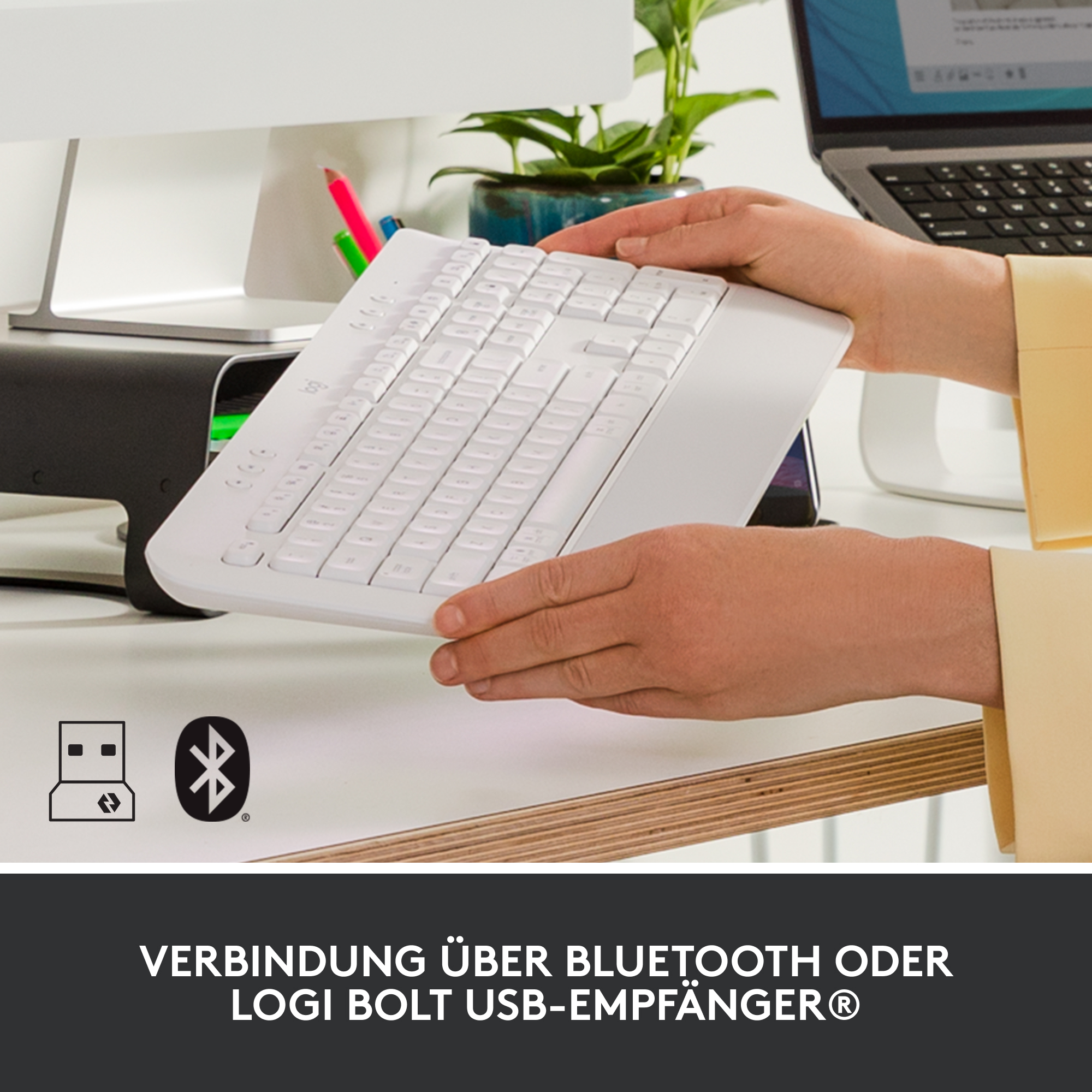 Weiße Tastatur mit USB- und Bluetooth-Symbolen auf dem Schreibtisch. Ein Laptop im Hintergrund.