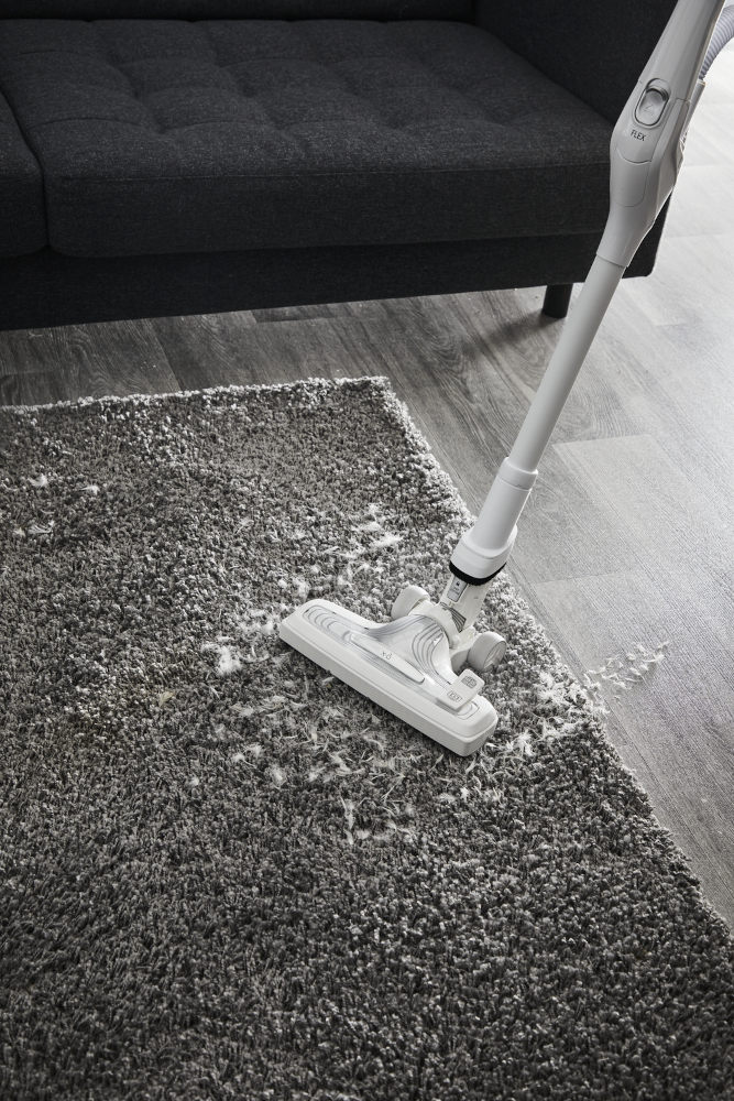 Een witte stofzuiger maakt een grijs tapijt schoon in de buurt van een donkergrijze bank, met een houten vloer op de achtergrond.