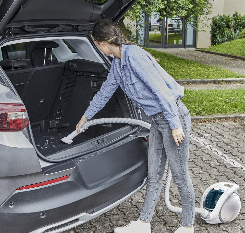 Een vrouw stofzuigt een kofferbak. Ze draagt een spijkerbroek en een blauw overhemd. Een stofzuiger staat op de stoep. De auto is grijs.