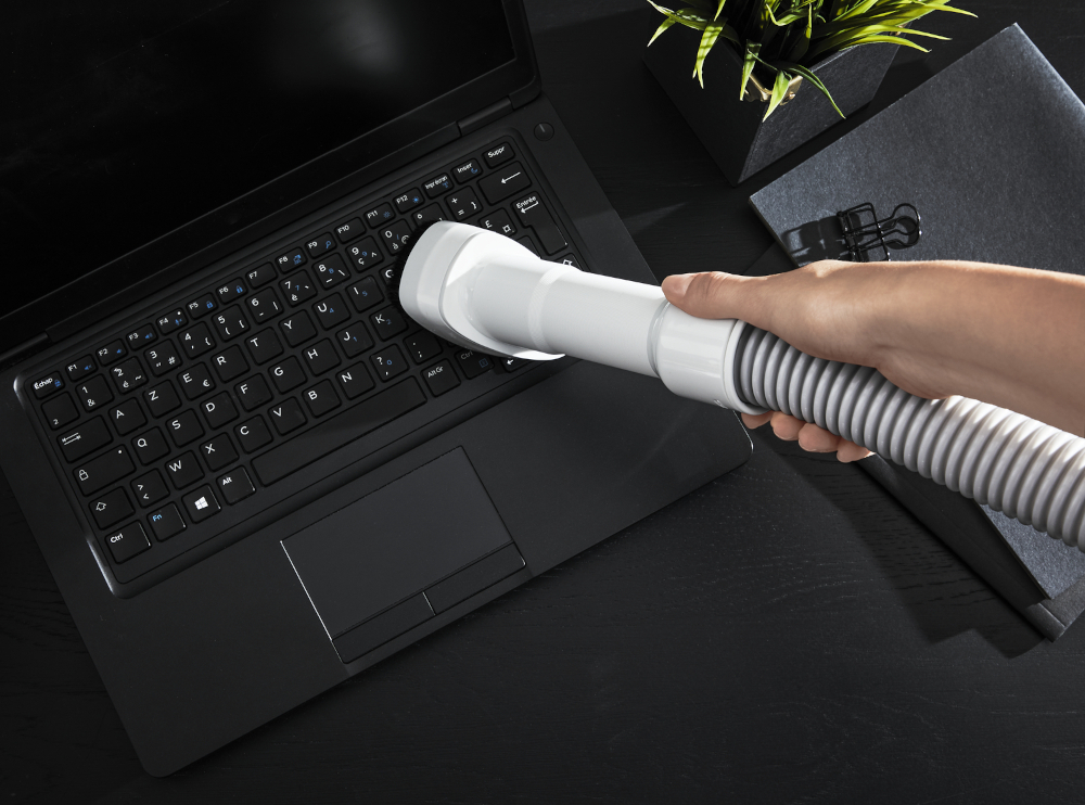 Een persoon maakt een laptoptoetsenbord schoon met een stofzuiger. Andere items op het zwarte bureau zijn een plant en een notitieboek.