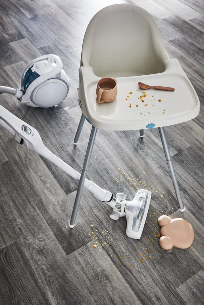 Een kinderstoel met eten op de plank, een gemorste vloeistof op de vloer en een stofzuiger die schoonmaakt.