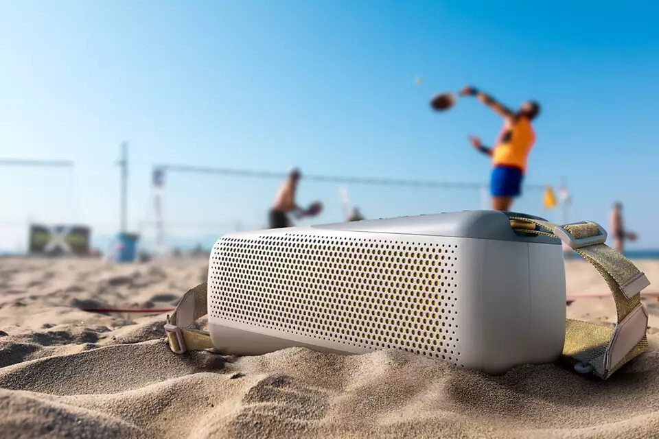 Biało-szary głośnik na piasku z ludźmi grającymi w siatkówkę plażową w tle.