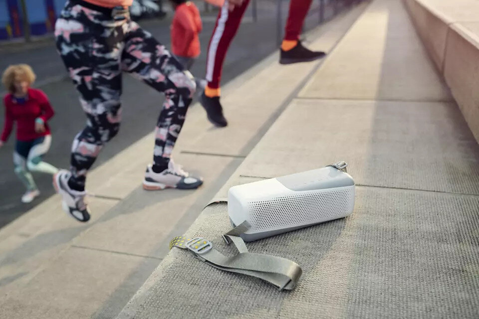 Biało-szary głośnik na schodach z ludźmi biegnącymi w tle. Głośnik ma pasek.