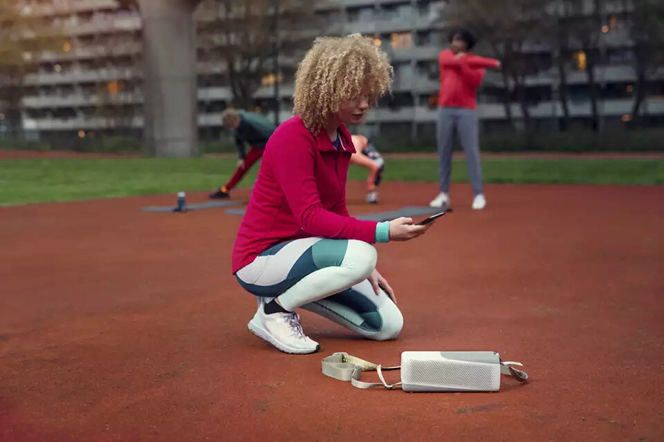 Kobieta używająca telefonu, kuca obok głośnika, a w tle ćwiczą inni ludzie.