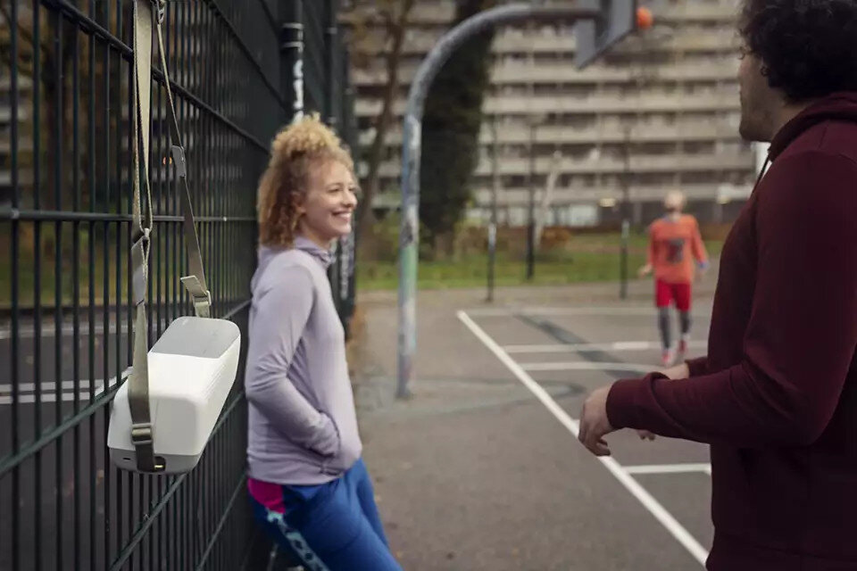 Kobieta oparta o ogrodzenie z wiszącym głośnikiem przenośnym i mężczyzną w tle.