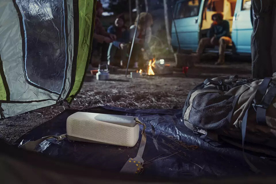 Scena kempingowa z głośnikiem, namiotem, ogniskiem i ludźmi.