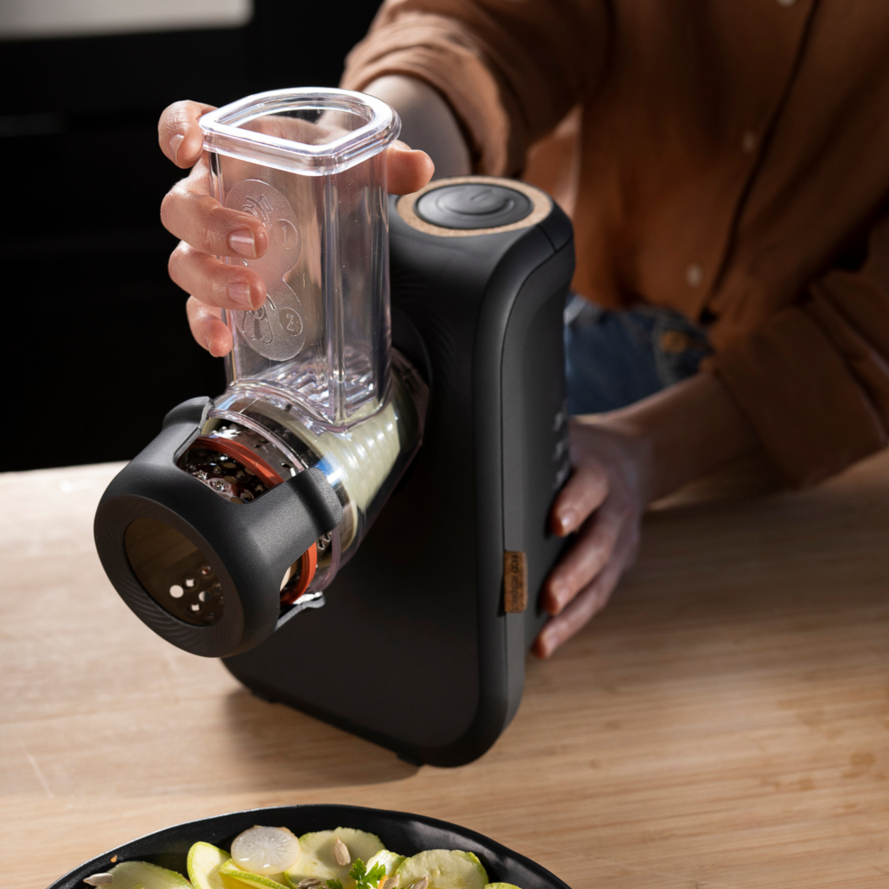 Une personne utilise un robot culinaire noir avec un récipient transparent pour râper des légumes.