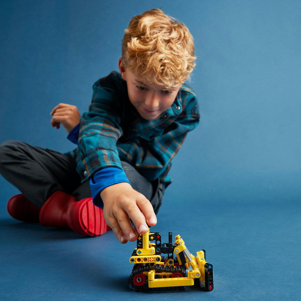 Ein Junge spielt mit einem gelben LEGO-Bagger-Modell auf einer blauen Oberfläche.
