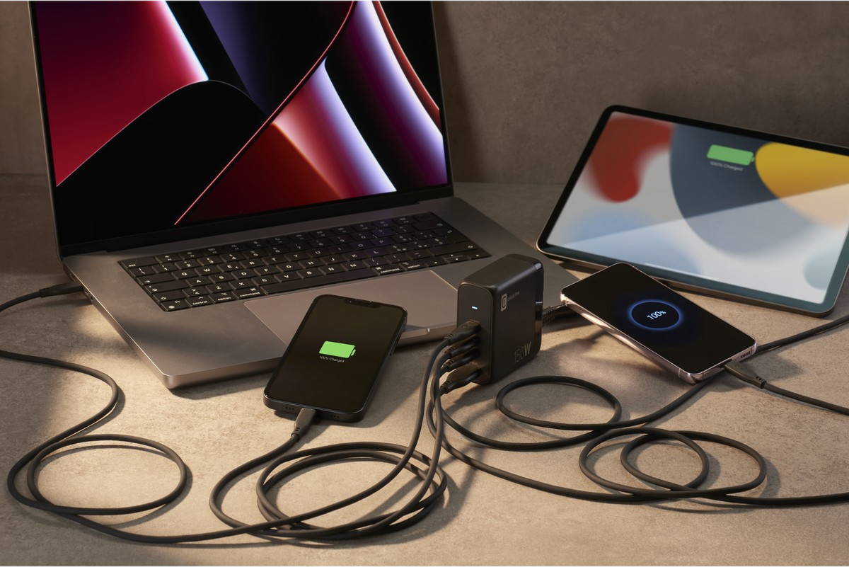 Mehrere Geräte laden an einem schwarzen Netzteil auf einem Schreibtisch. Geräte wie Laptop und Telefone.
