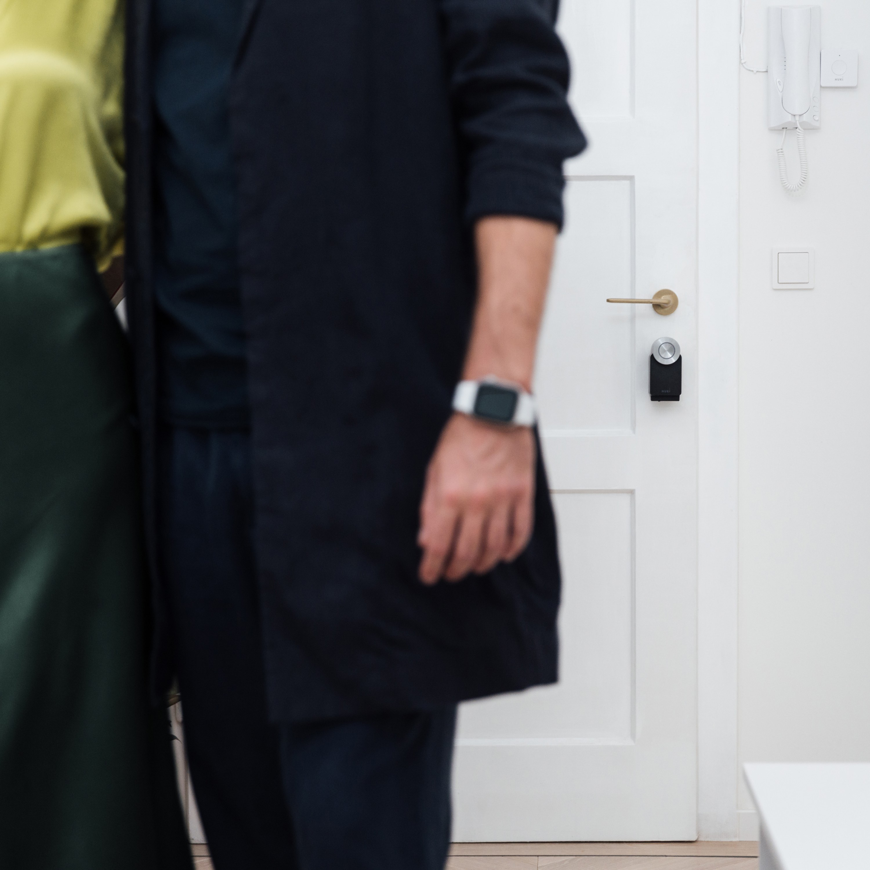 Una coppia è in piedi vicino a una porta bianca con una serratura intelligente nera. Si vede un orologio al polso dell'uomo.