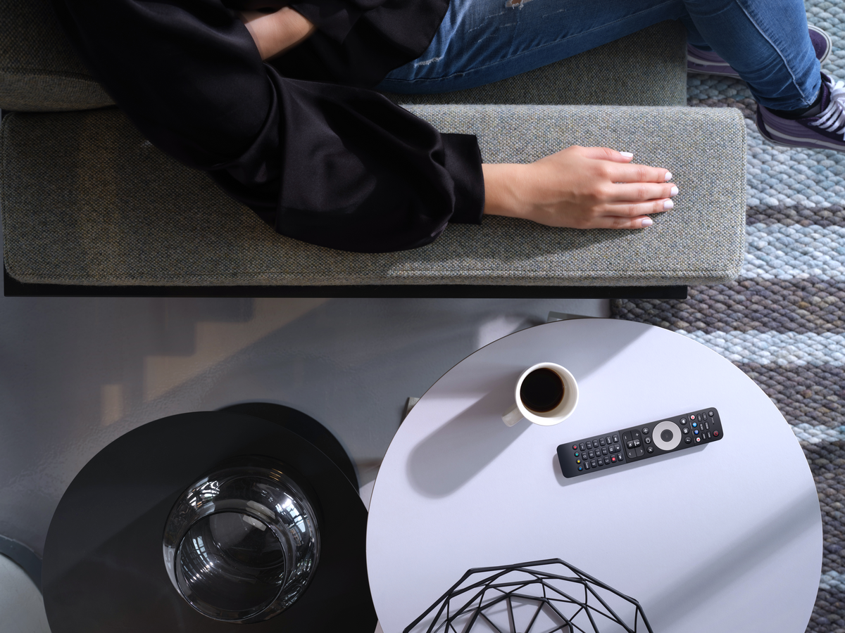 Person auf Couch, Hand ruht. Kaffee, Fernbedienung auf weißem Tisch. Glasvase auf schwarzem Tisch. Teppich, Treppe im Hintergrund.