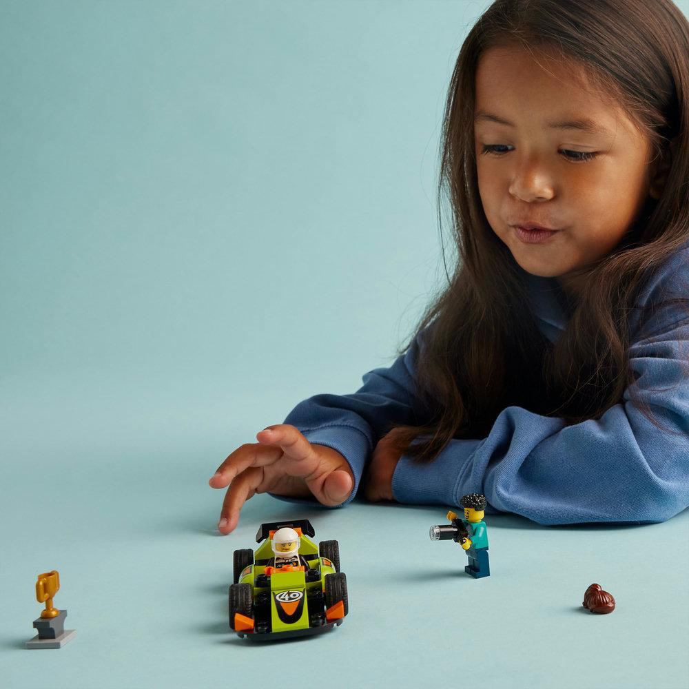 Ein Mädchen spielt mit Lego-Rennwagen und Figuren. Eine Trophäe und ein braunes Objekt liegen auf dem blauen Tisch.