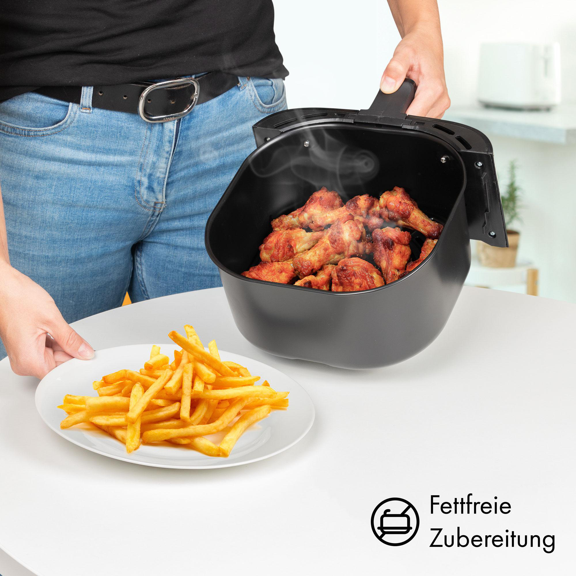 Eine Person hält eine Heißluftfritteuse mit gekochten Hähnchenflügeln. Pommes frites sind auf einem weißen Teller.