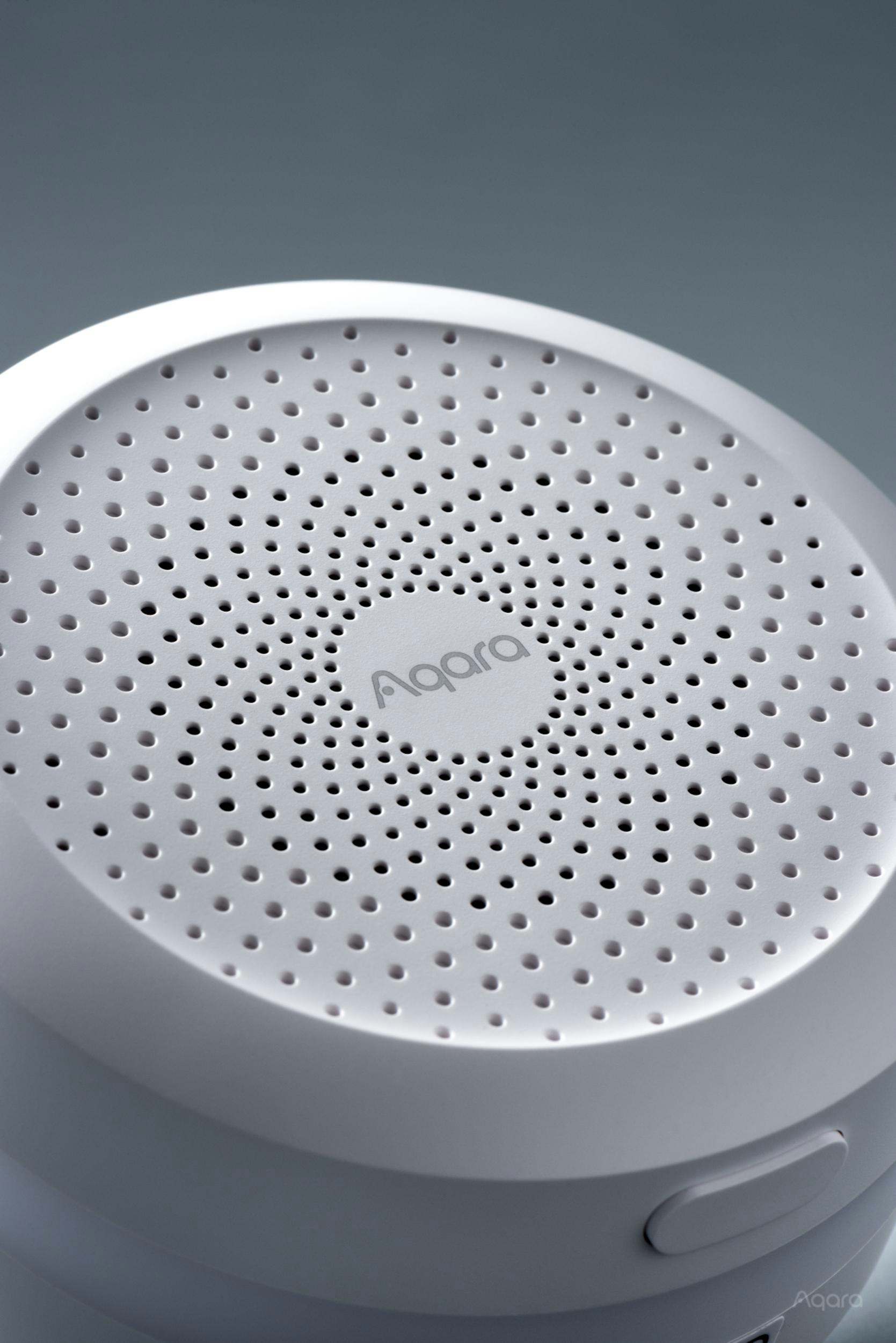 Ein weißes, rundes Aqara Smart-Home-Gerät mit einem Gitter aus kleinen Löchern und dem Markenlogo in der Mitte.