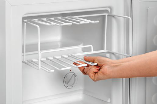 Eine Hand verstellt ein weißes Drahtregal in einem weißen Kühlschrank. Ein Bedienknopf ist sichtbar. Das Innere des Kühlschranks ist weiß.