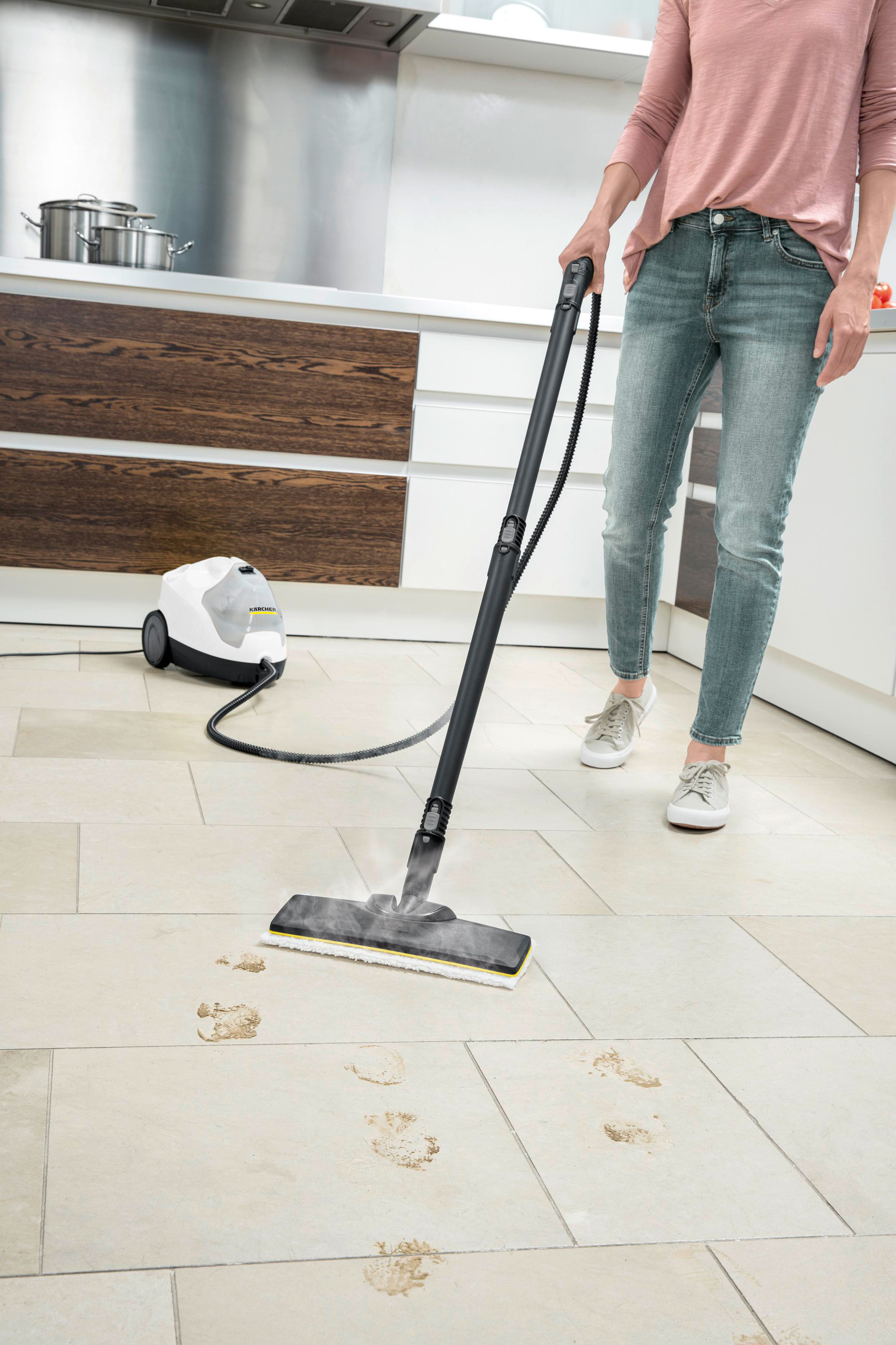 Frau reinigt den Boden mit einem Dampfreiniger. Der Boden hat Fußabdrücke. Im Hintergrund sind ein Dampfreiniger und eine Küche.