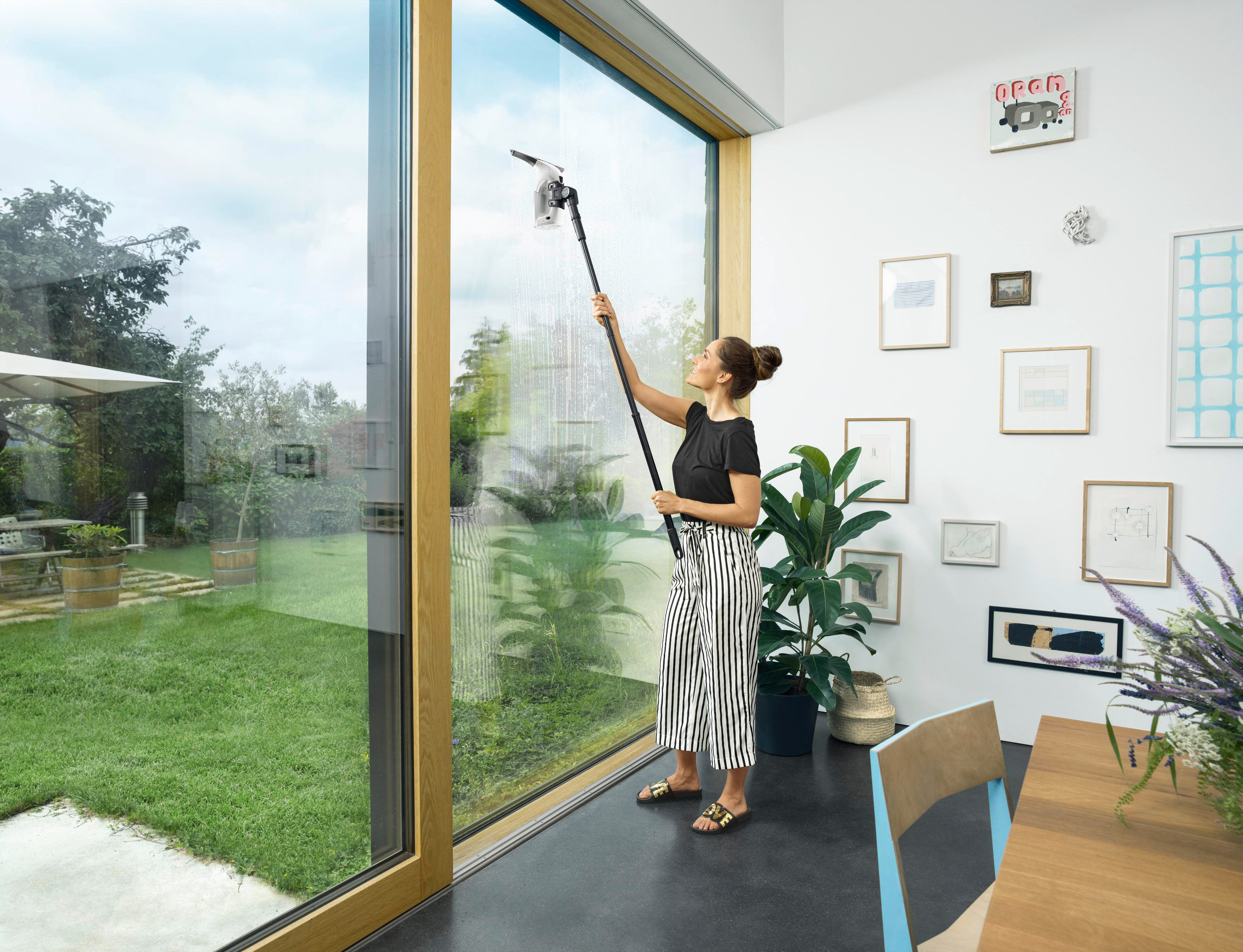 Eine Frau reinigt ein Fenster mit einem Langstielreiniger. Ein Garten und Innenraum sind sichtbar.