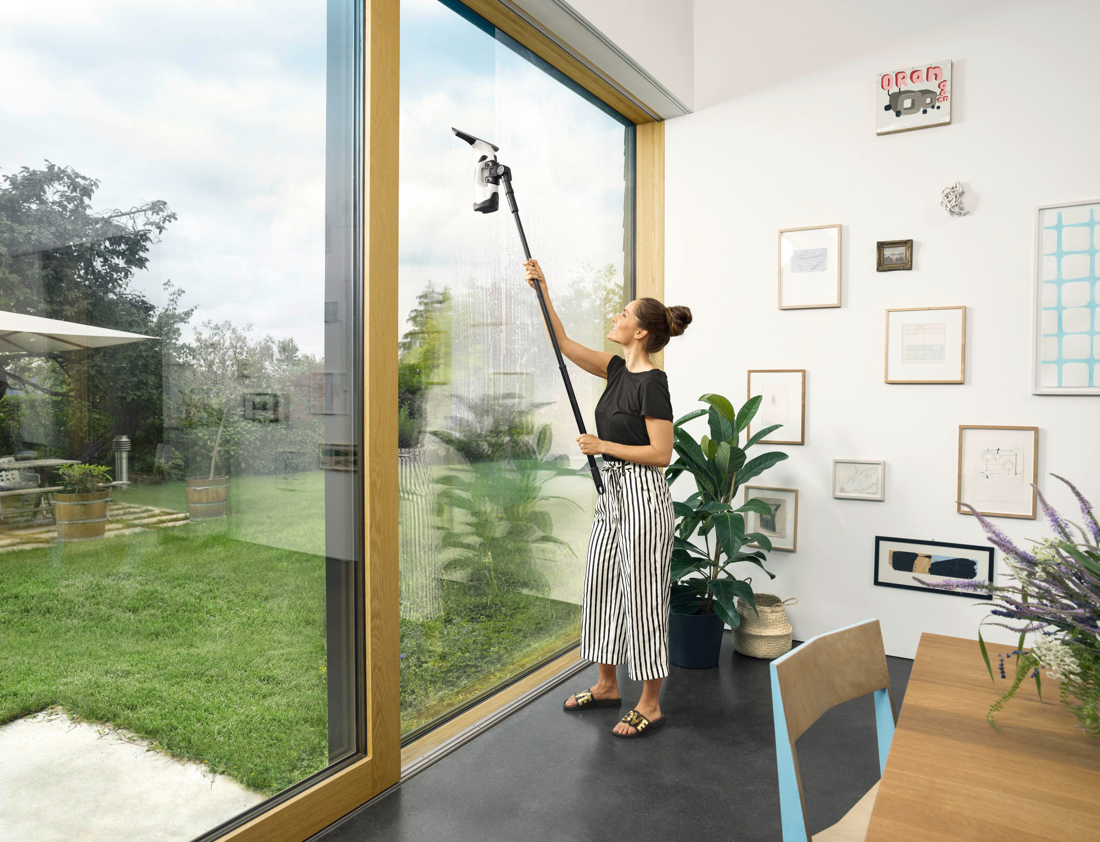 Eine Frau reinigt ein Fenster mit einem Fensterreiniger mit langem Griff, im Hintergrund ein Garten.