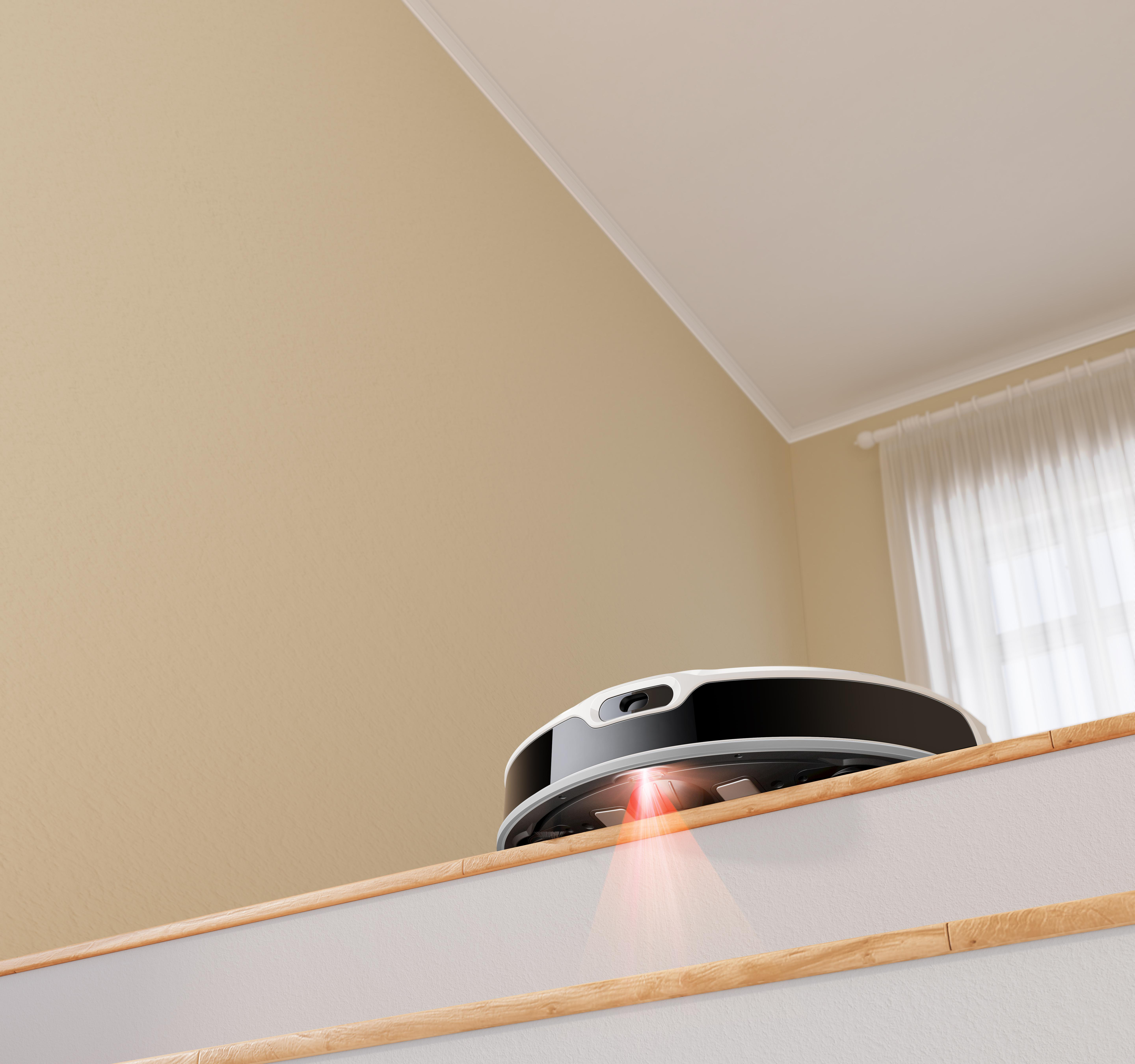 Roboter-Staubsauger auf einer Treppe. Beige Wand und weiße Decke mit Fenster und Vorhang.