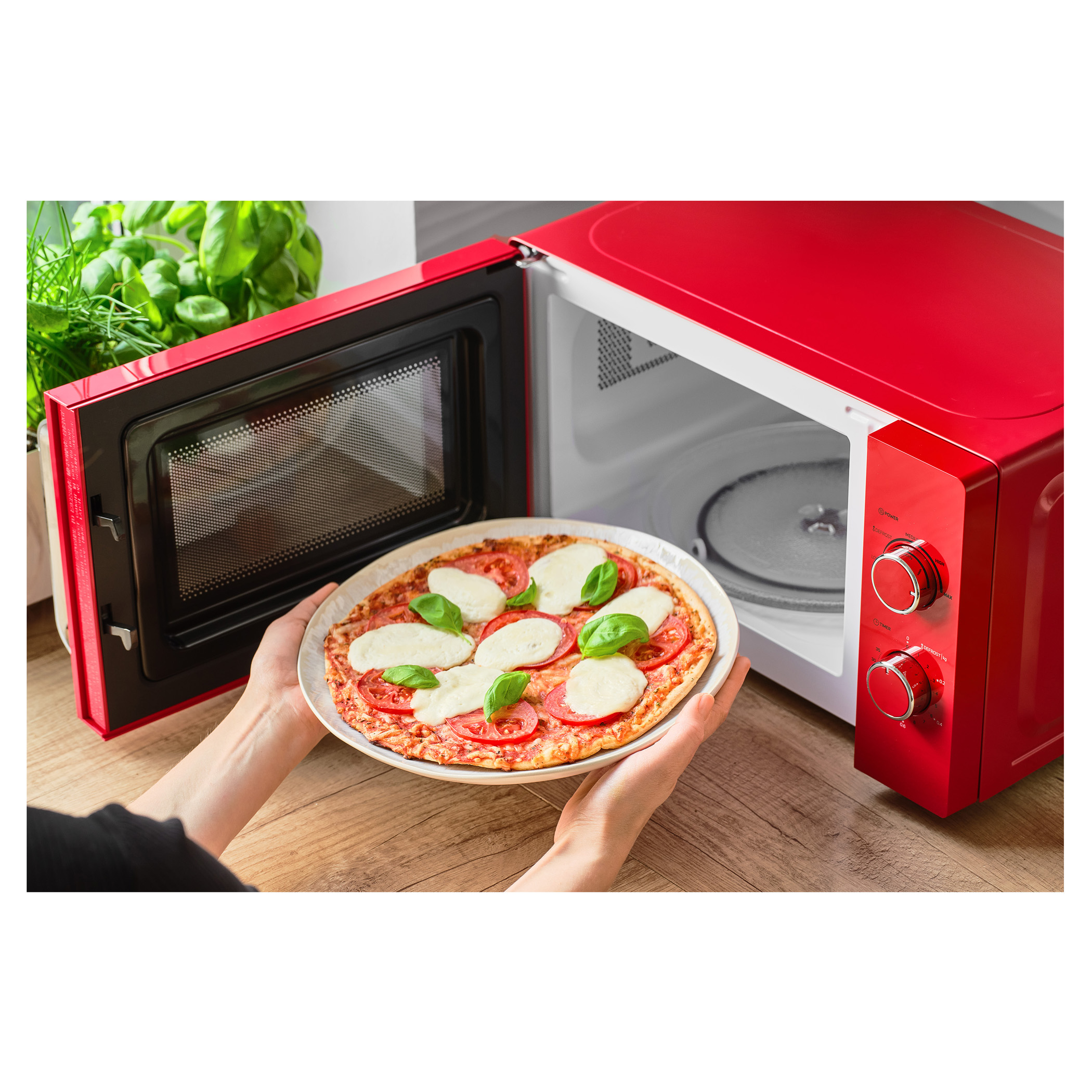 Egy piros mikrohullámú sütő nyitott ajtóval, egy tányéron pizzát mutat, amelyet két kéz vesz ki.