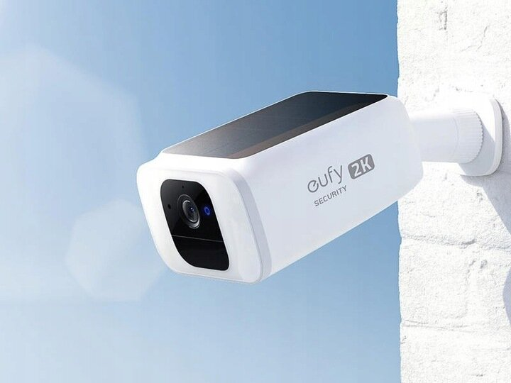 Biała kamera monitoringu zamontowana na białej ceglanej ścianie, z błękitnym niebem.