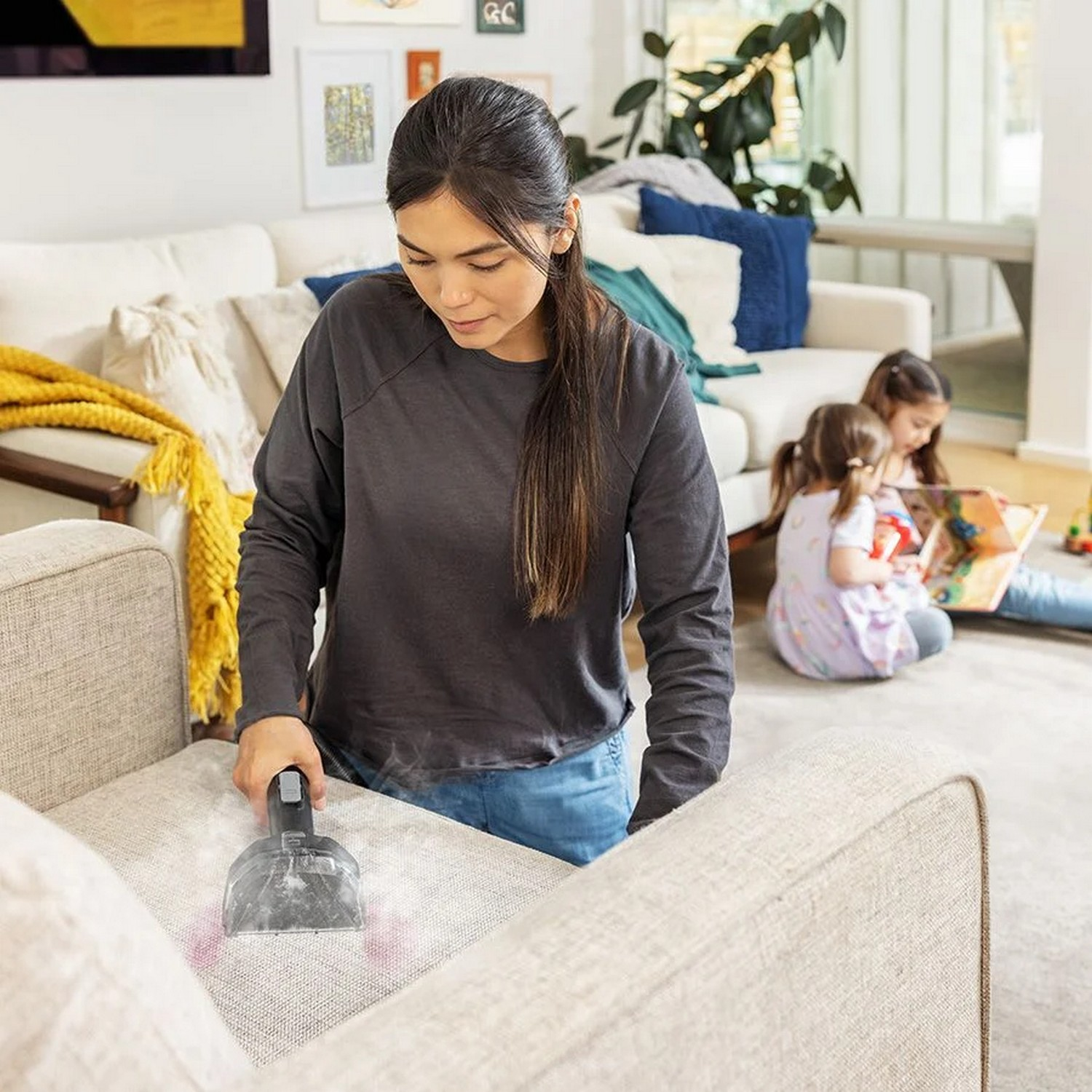 Woman vacuuming a couch, two children reading a book in the background.