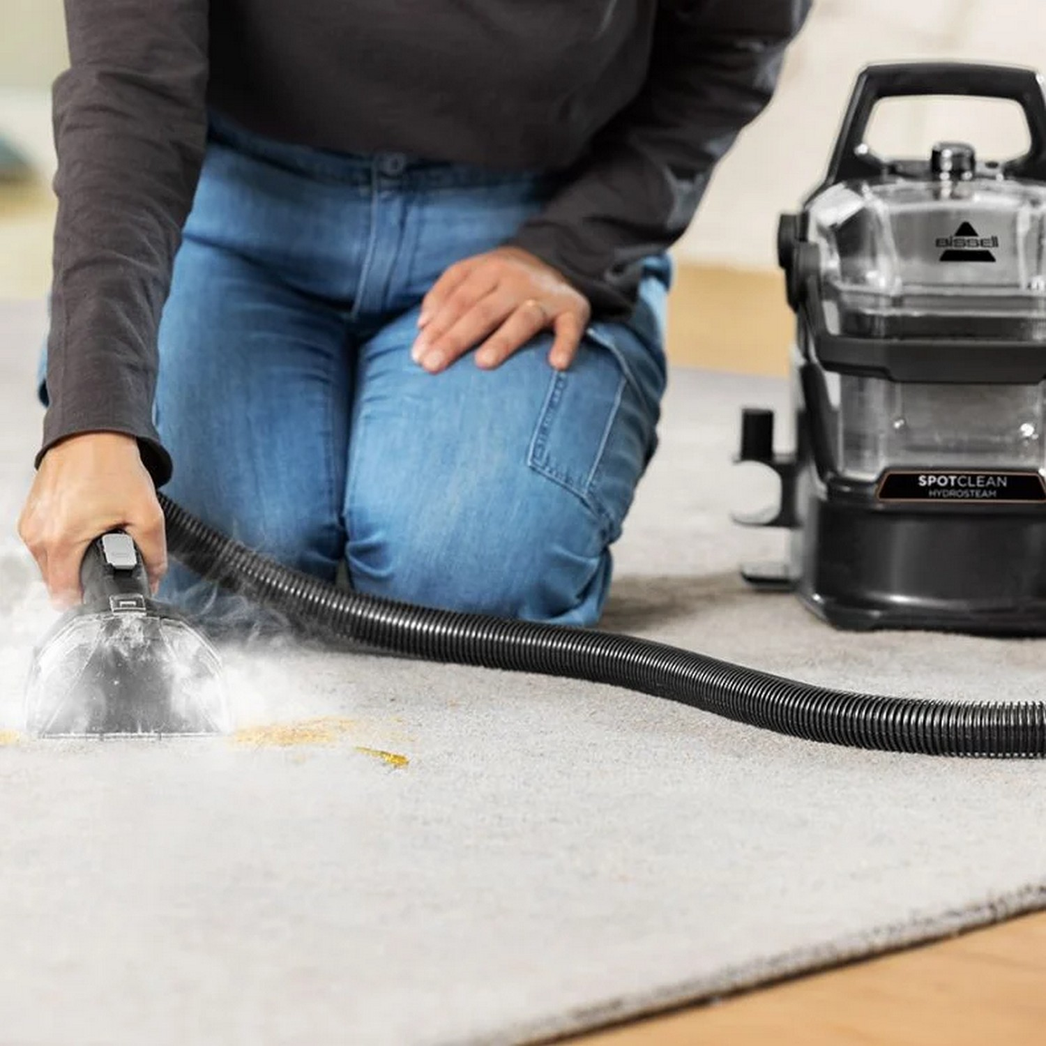 A person using a spot cleaner to remove a stain from a gray carpet. The cleaner is black.