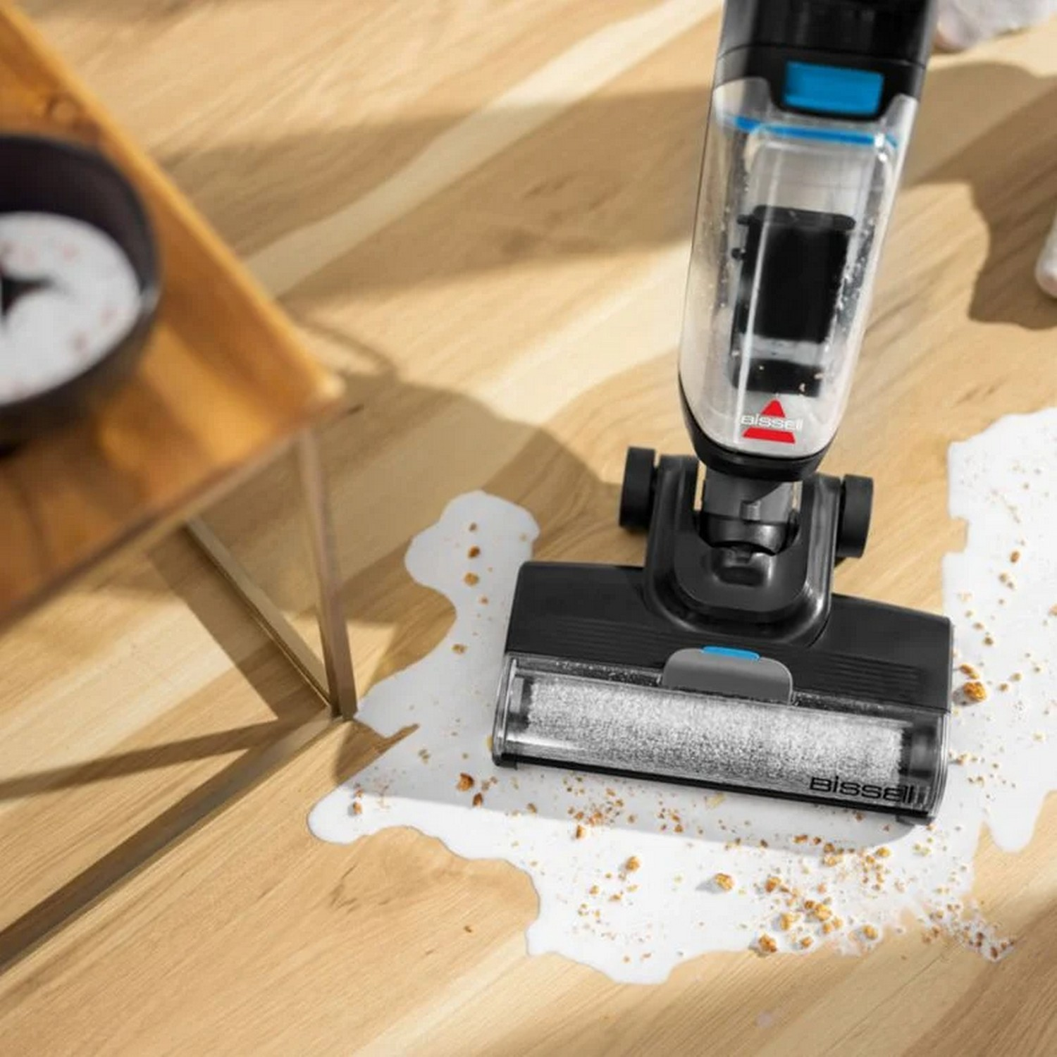 Un aspirateur Bissell nettoie un déversement sur un plancher en bois. De l'eau et des débris sont visibles.