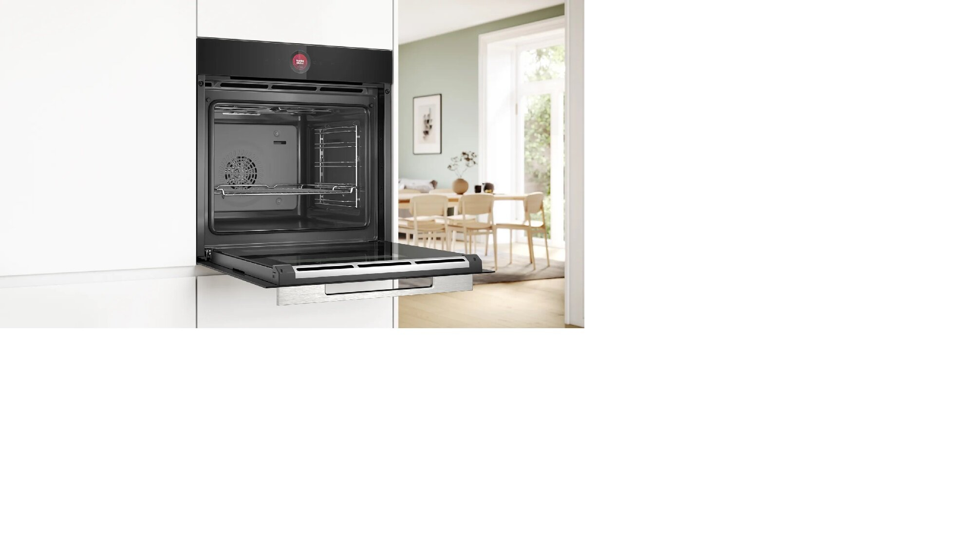 Otwarty piekarnik w zabudowie w białej kuchni. Drzwi piekarnika są otwarte. W tle jadalnia.