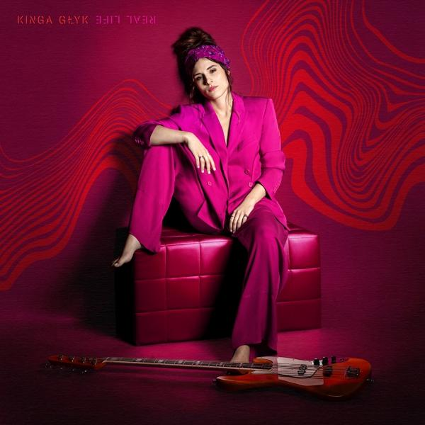 Rózsaszín öltönyös nő piros kockán ül, basszusgitár előtt. Piros háttér hullámzó vonalakkal. 'Real Life' album borítója.