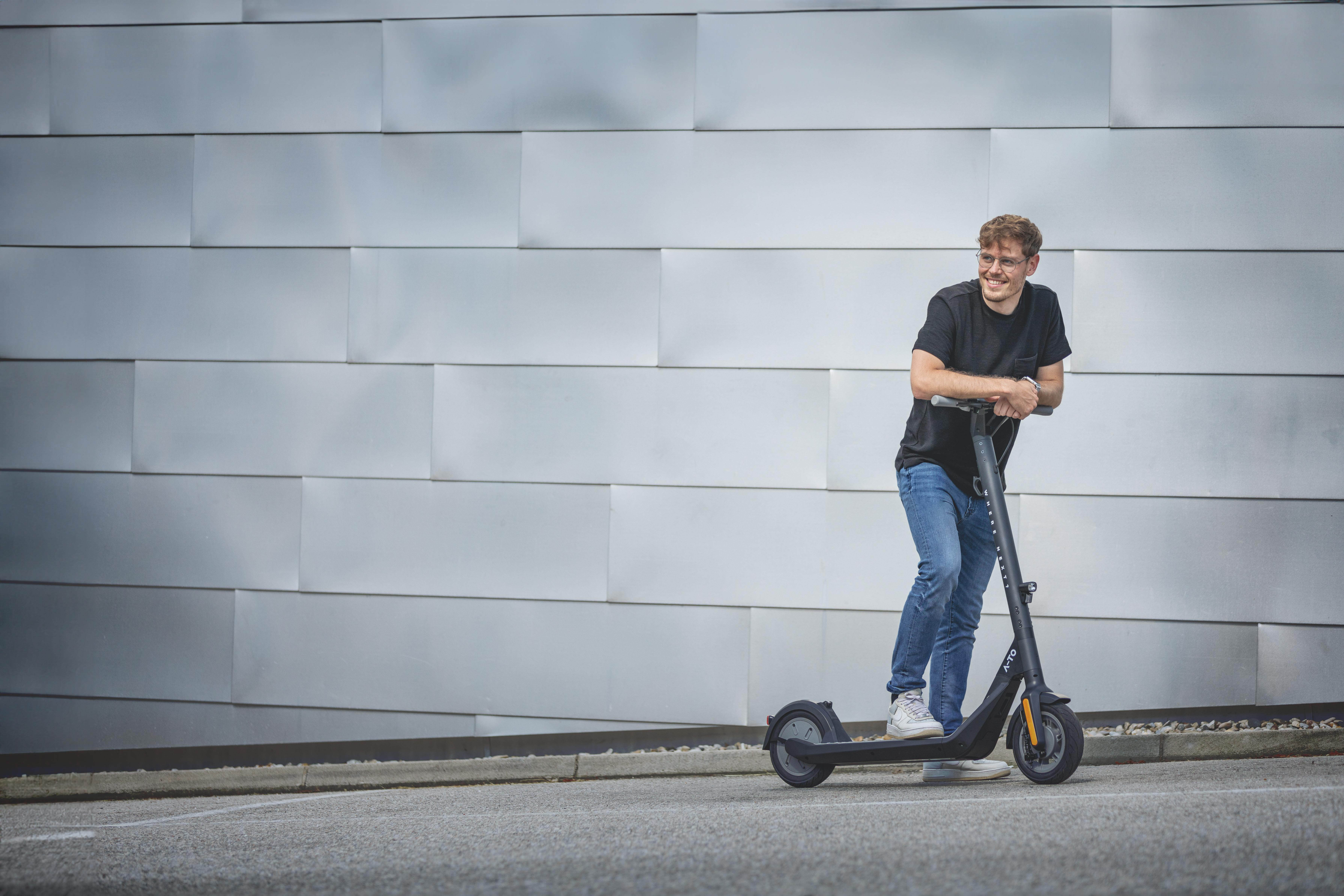 Ein Mann auf einem Roller lächelt, lehnt sich an den Lenker. Hintergrund ist eine silberne Wand.