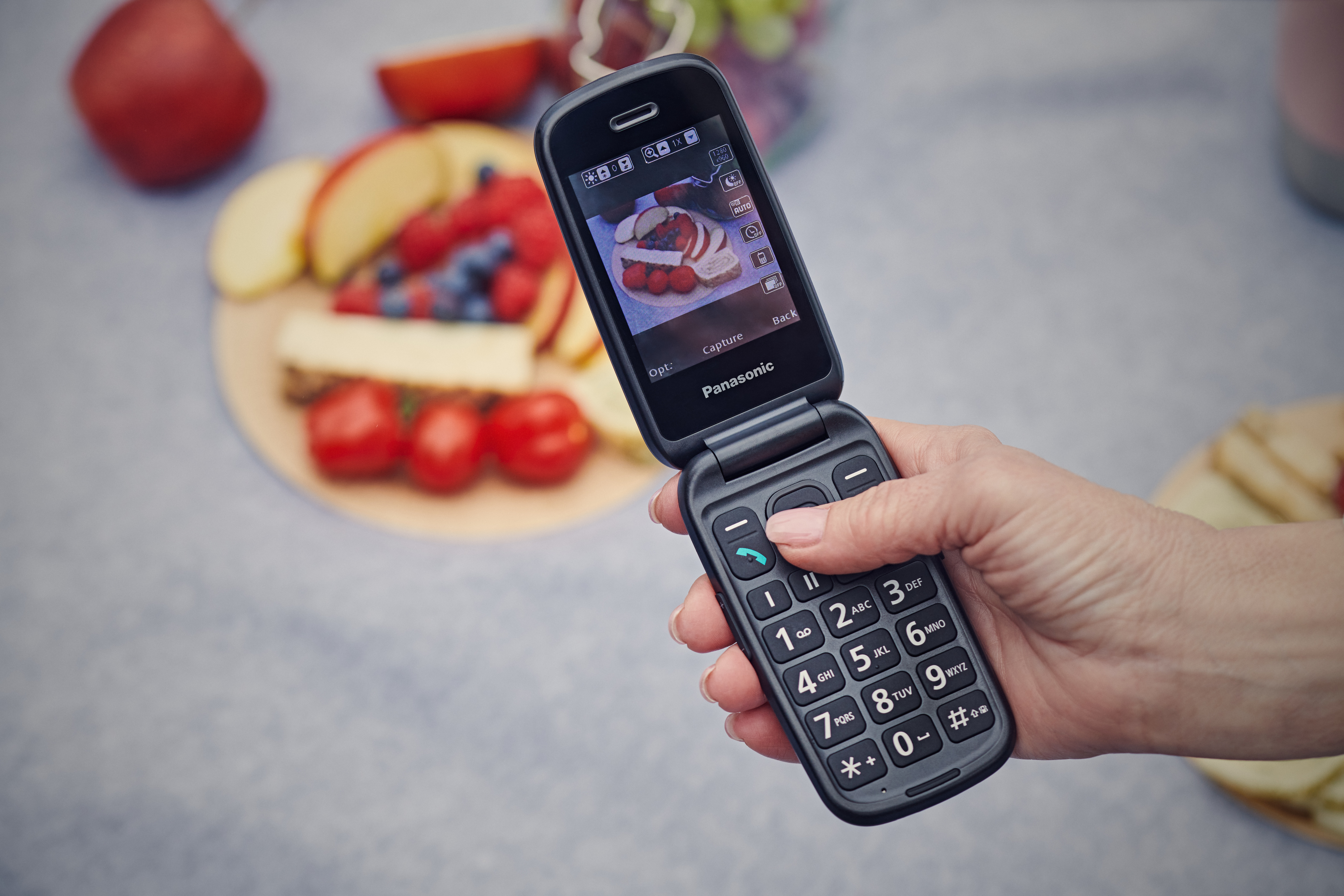 Mano sosteniendo teléfono plegable, pantalla mostrando comida. Plato con fruta y comida al fondo.