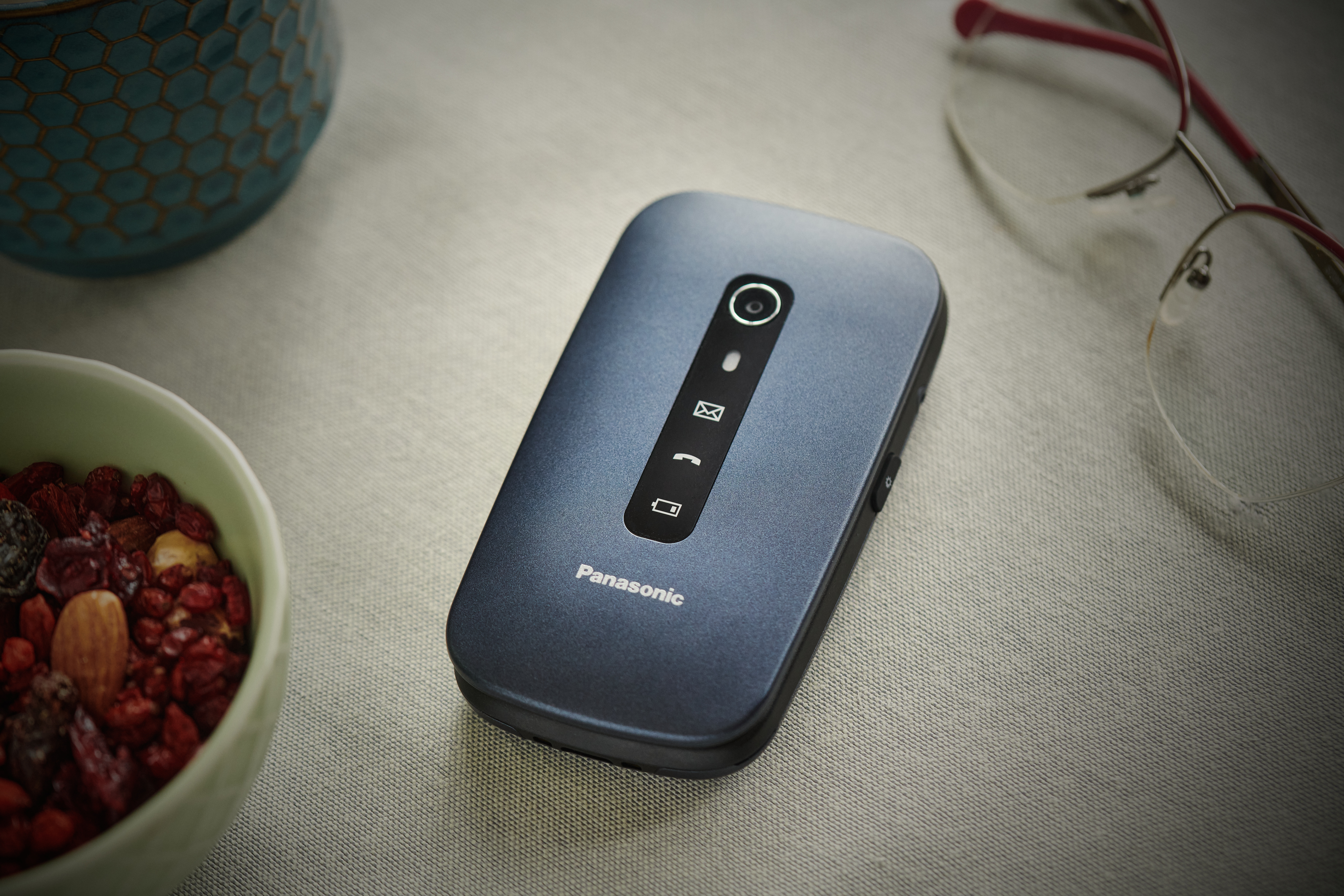 Un teléfono plegable azul está sobre una superficie blanca. Junto a él, un cuenco de frutos secos y gafas.
